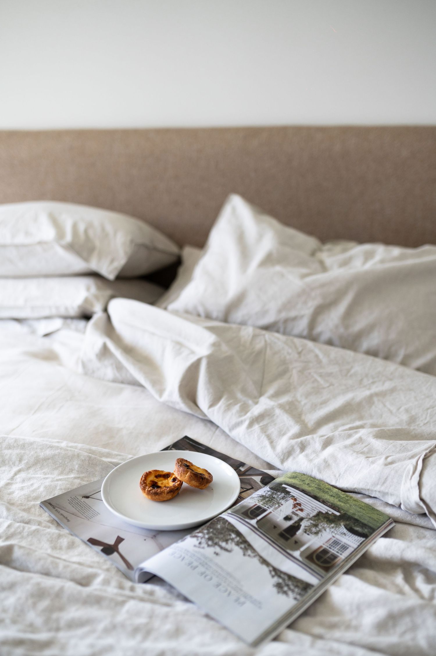 A series of minimalist photos showing white rumpled bedding with magazines and a small cookie on a plate.