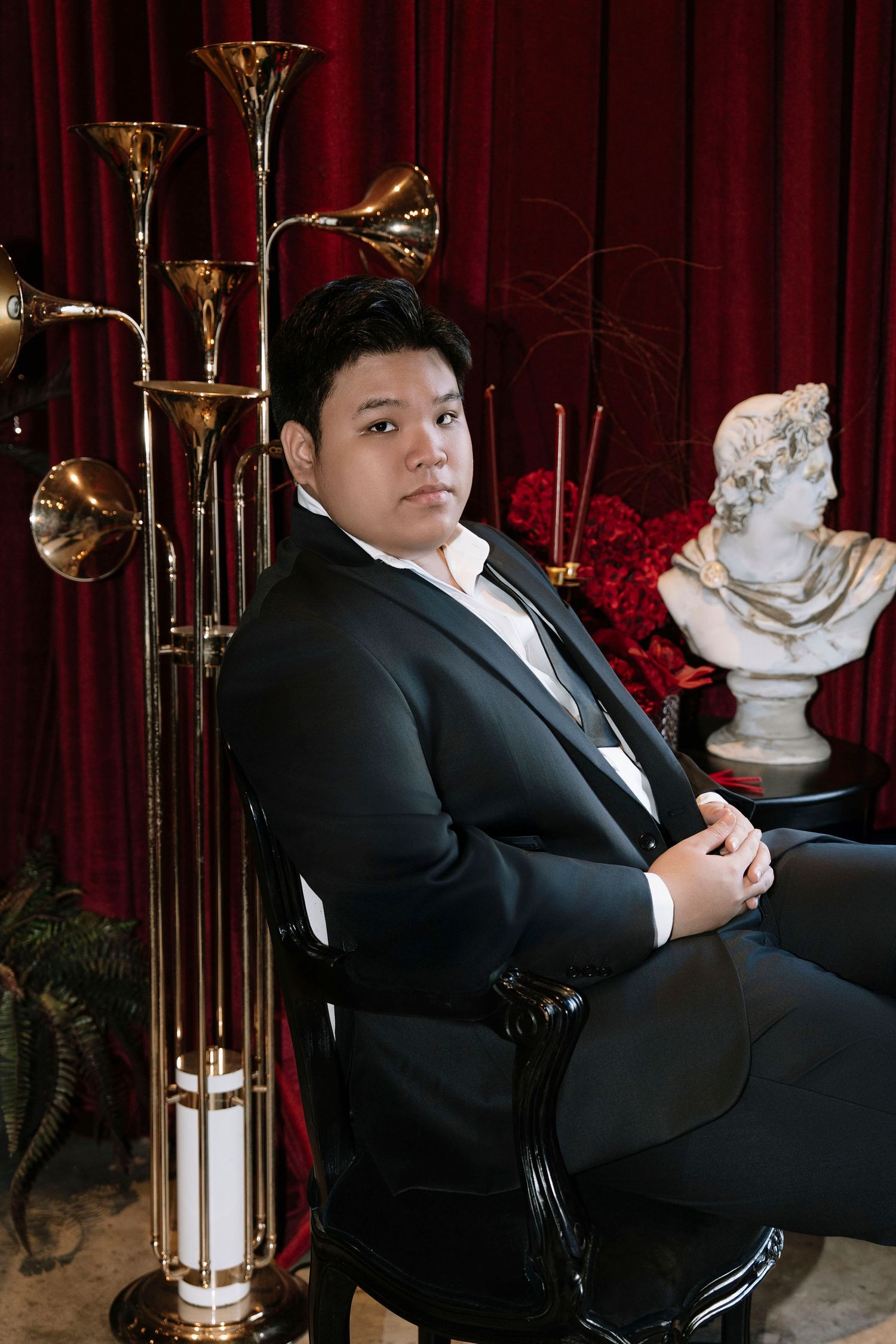 Asian groom in black suit seated in ornate chair beside gold floor lamp and marble bust sculpture.