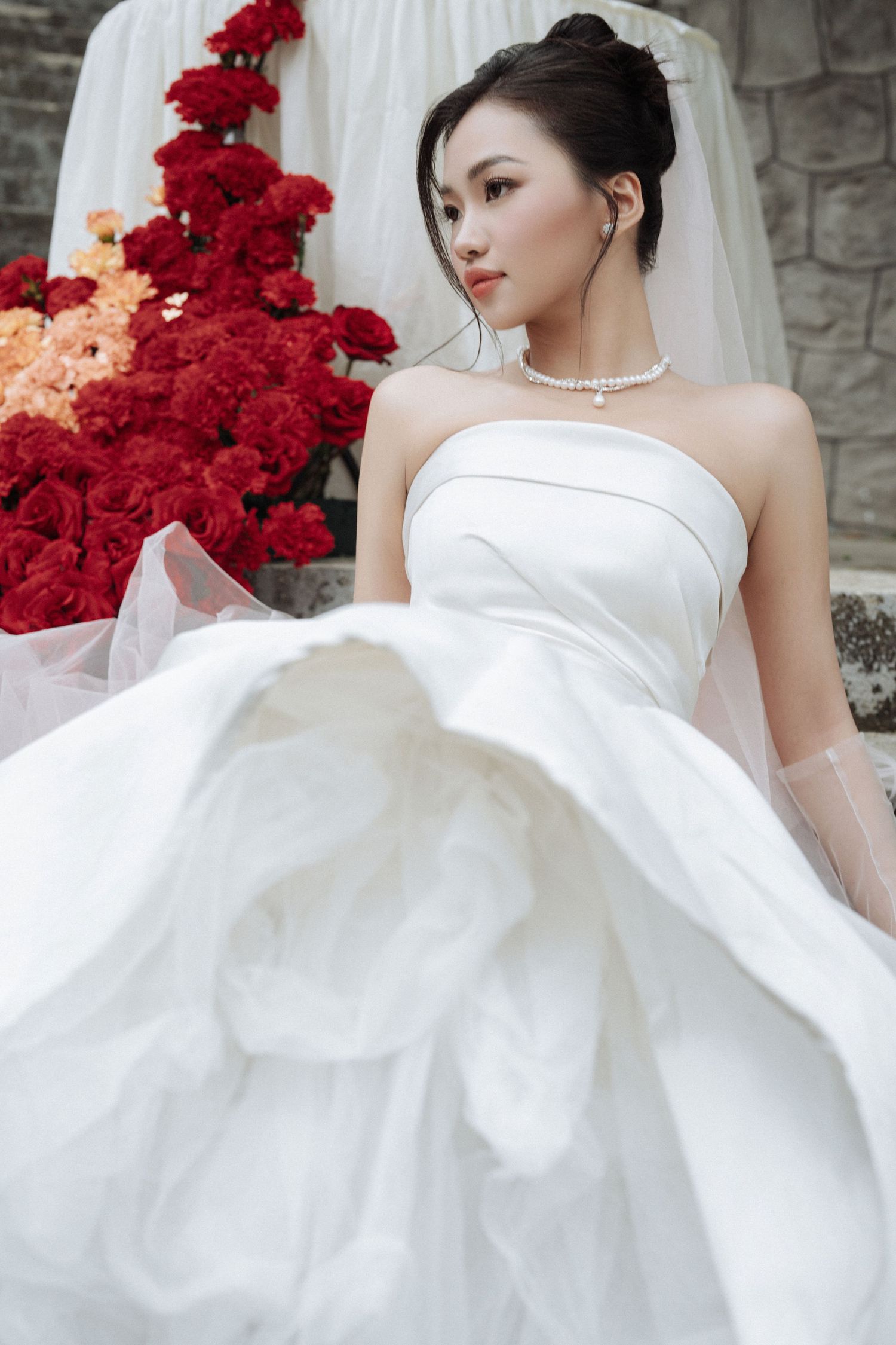 Elegant Asian bride in strapless white ball gown with pearl necklace and veil, seated near red floral arrangement.