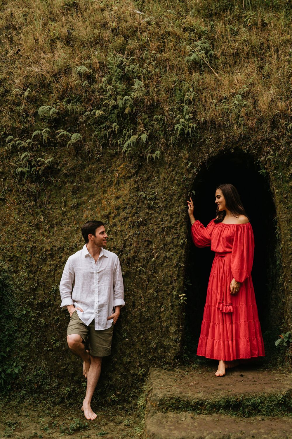 A couple in white and red attire pose playfully against an earthen wall in a natural setting.