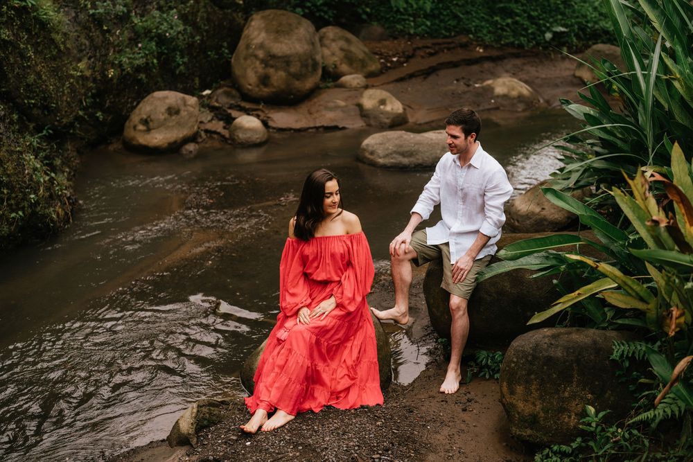 Couple enjoying moment together by rocky stream in tropical setting wearing red dress and white shirt.