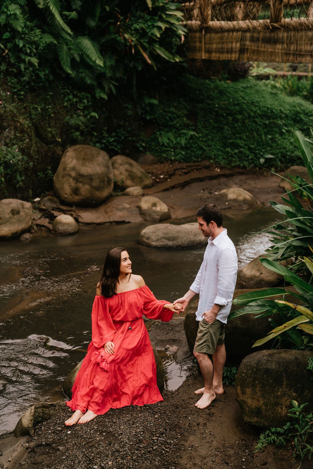 Two people relax by a stream with rocks and tropical foliage in the background.