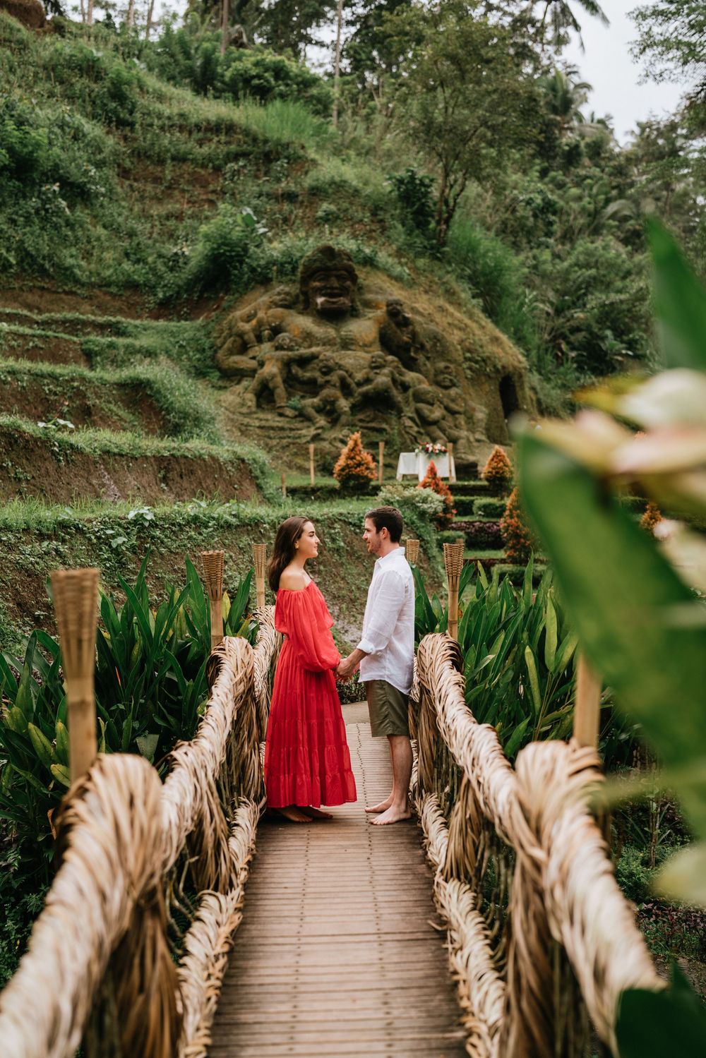 A couple walks along a wooden bridge decorated with dried pampas grass in a lush tropical setting.