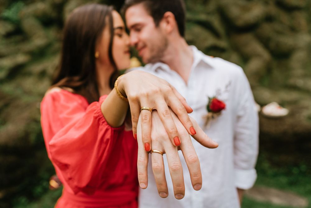 Close up of hand displaying engagement ring with couple embracing in background.