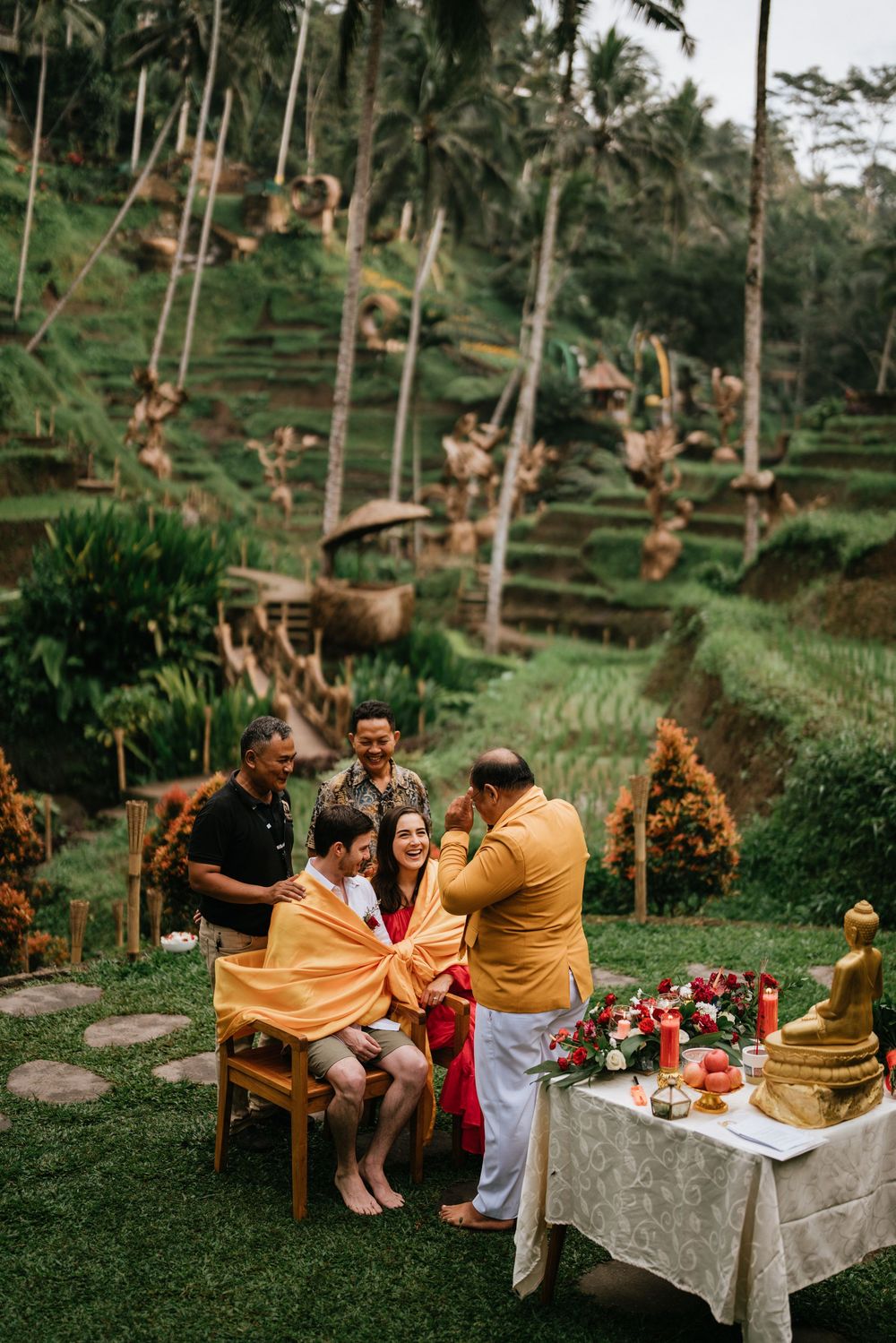 Intimate outdoor ceremony with guests gathered around decorated table in tropical rice terrace setting.