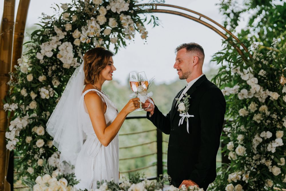 Couple exchanging vows under an arch adorned with white flowers and greenery.