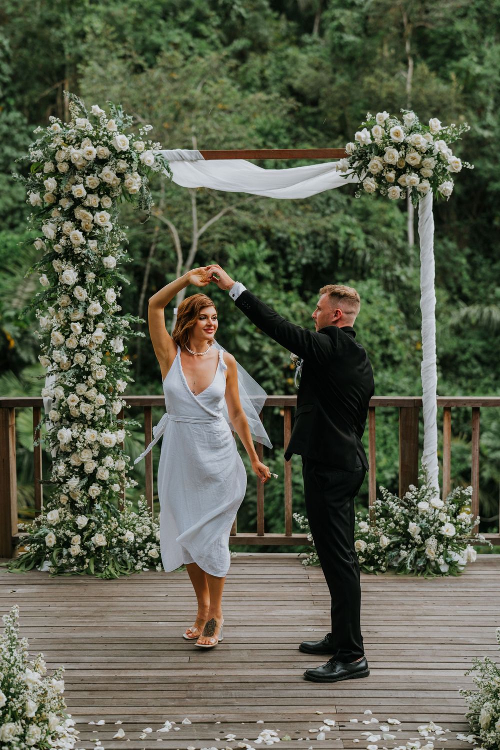Playful wedding moment on wooden deck decorated with white roses and scattered petals.