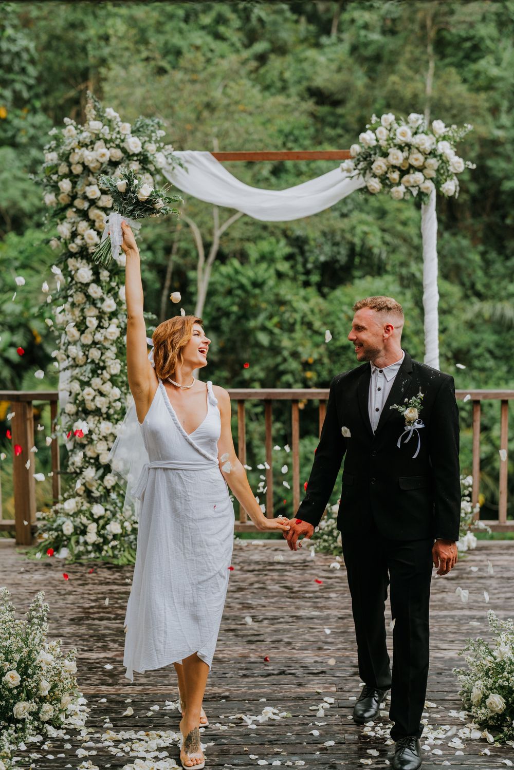 Celebratory wedding dance on petal-covered deck with floral arch backdrop.