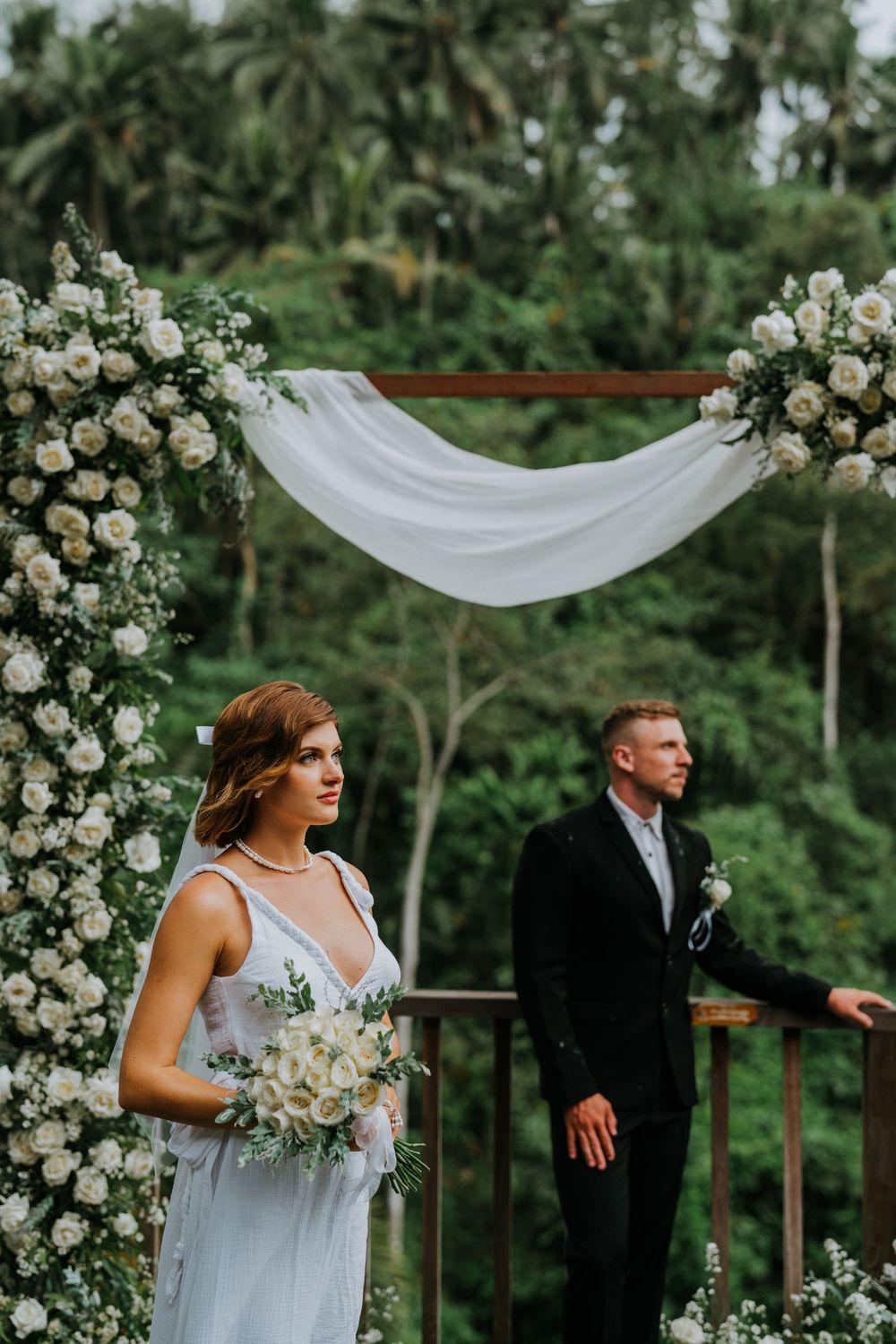 Elegant outdoor wedding ceremony setup with white floral arch and flowing drapery against a lush green forest backdrop.