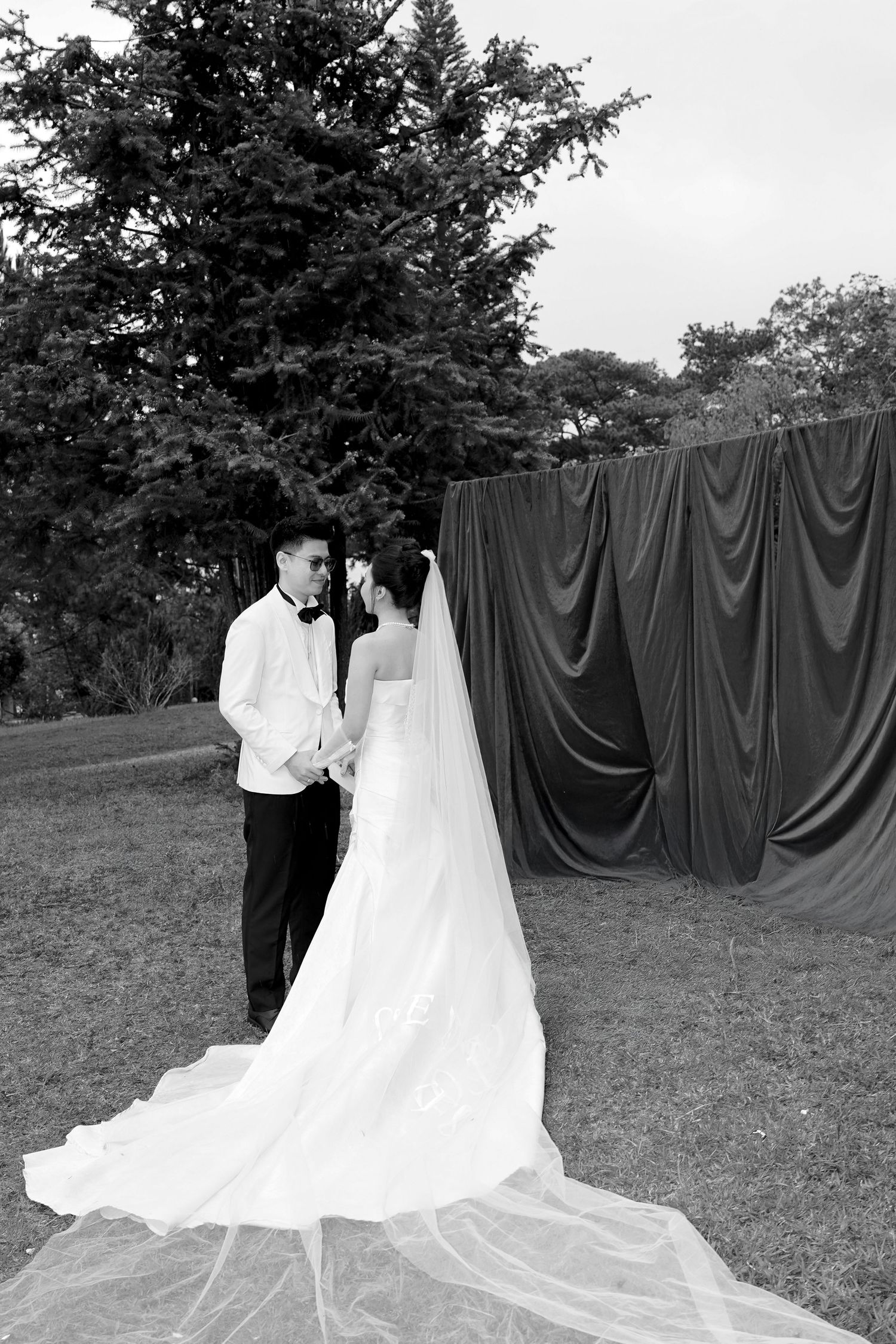 Black and white photo of bride and groom holding hands before draped fabric backdrop in garden.