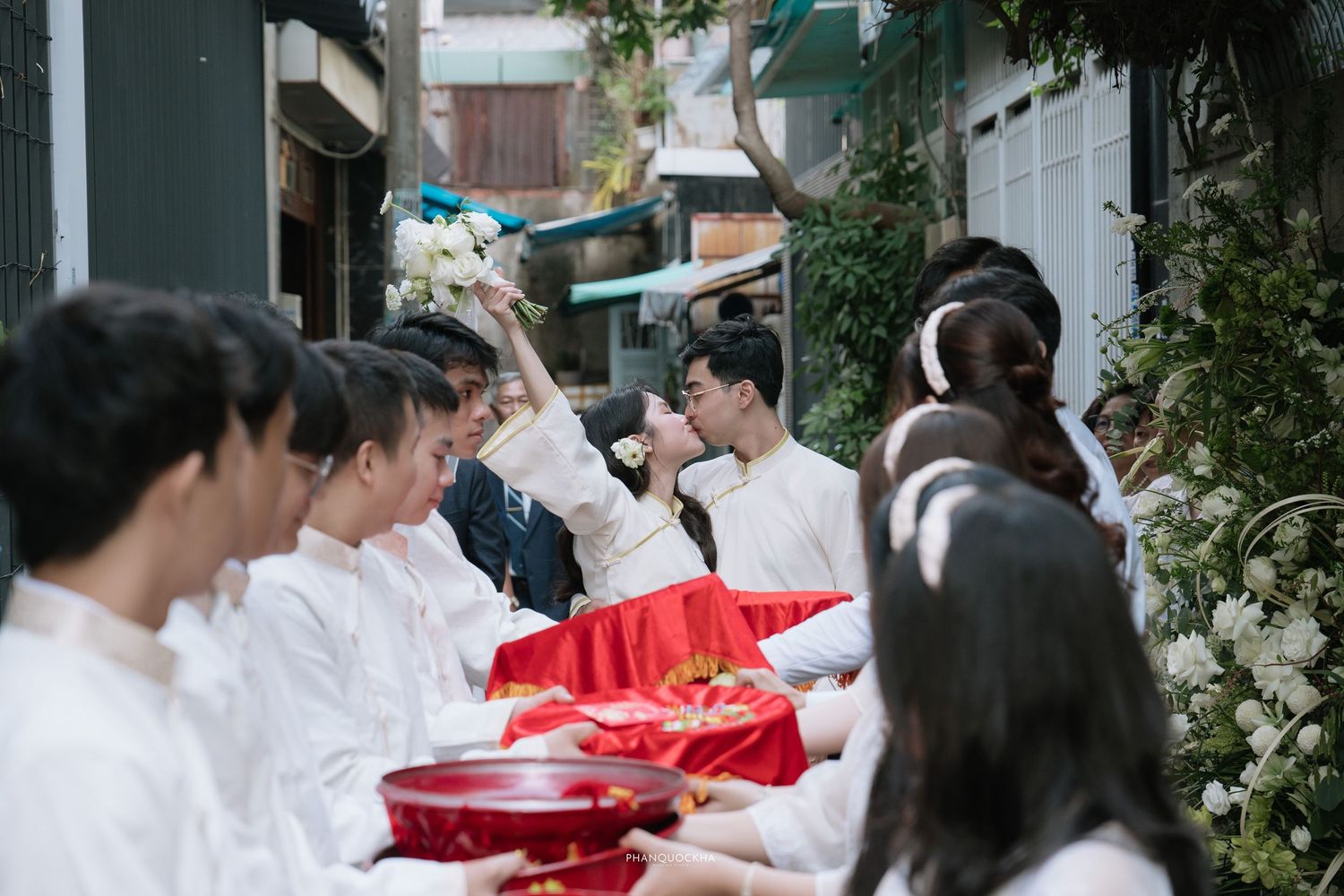 Vietnamese bride and groom kissing during traditional wedding ceremony with white-clad attendants nearby.