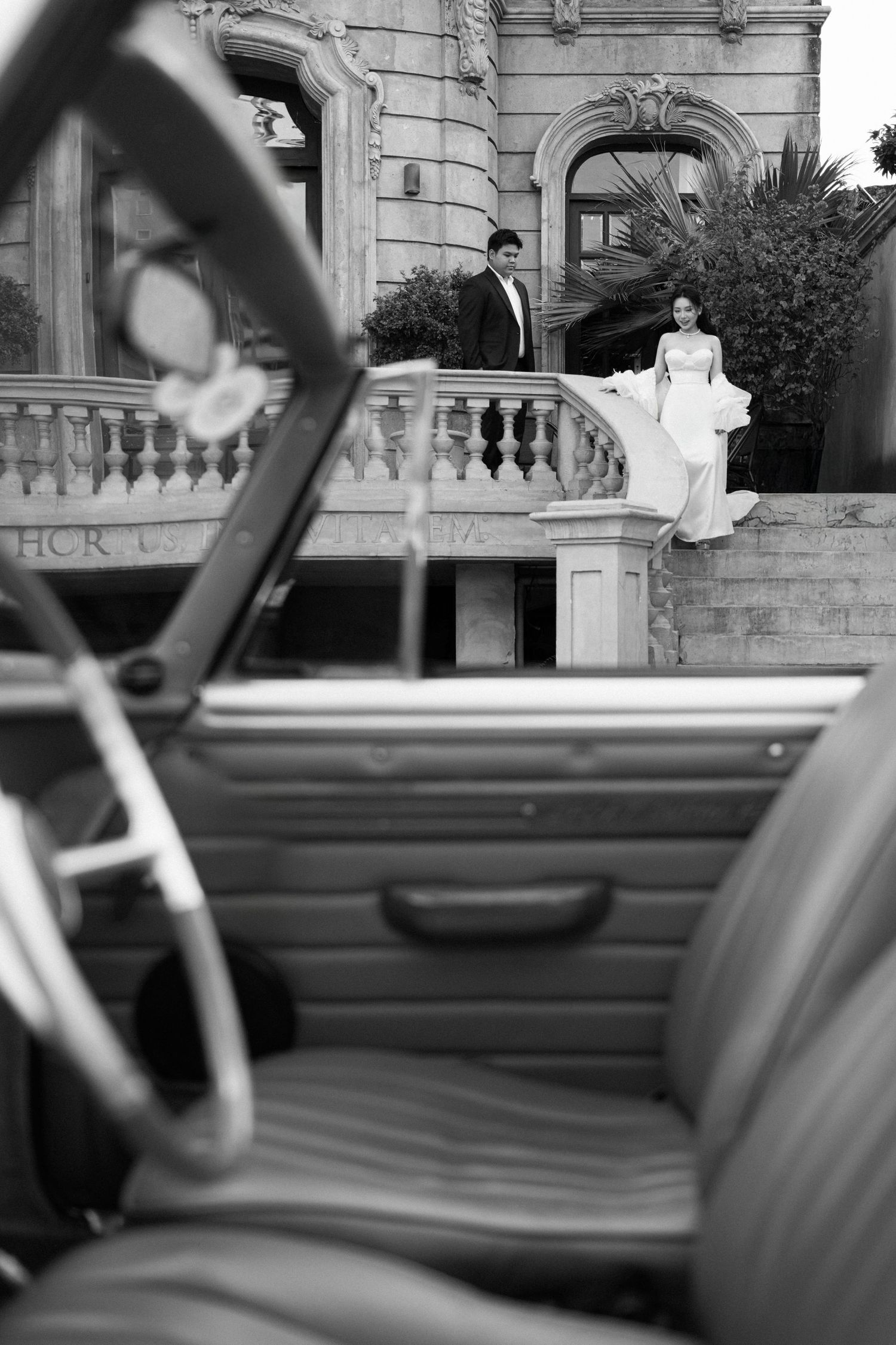 Black and white wedding photo of bride in white gown seated on balustrade with groom standing nearby, vintage car in foreground.