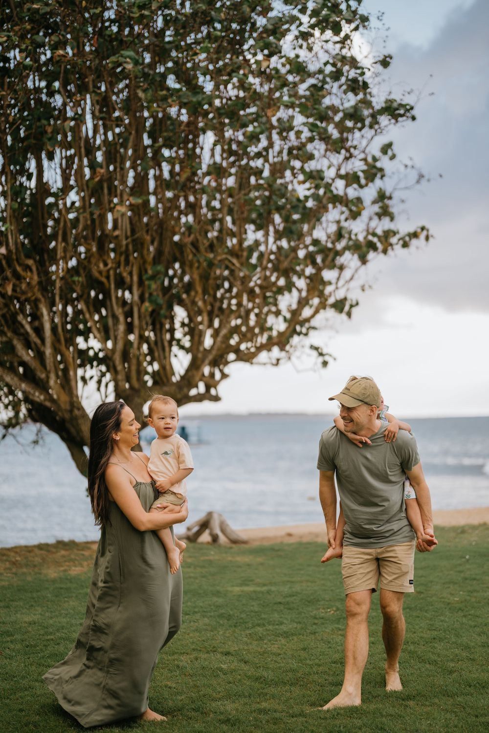 Family playing near a large tree by the ocean with cloudy skies overhead.