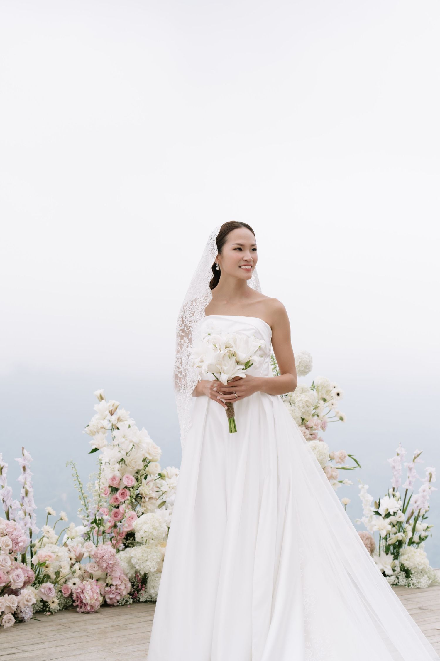 Smiling bride in white strapless gown holding bouquet stands before pink and white floral arch overlooking sea.