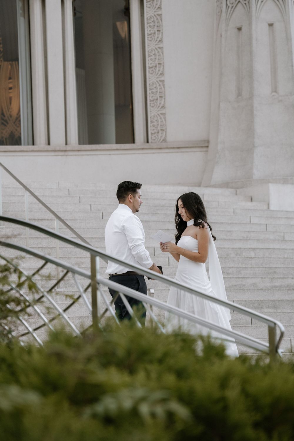 A couple in wedding attire poses on white marble steps with architectural columns and greenery in the foreground.