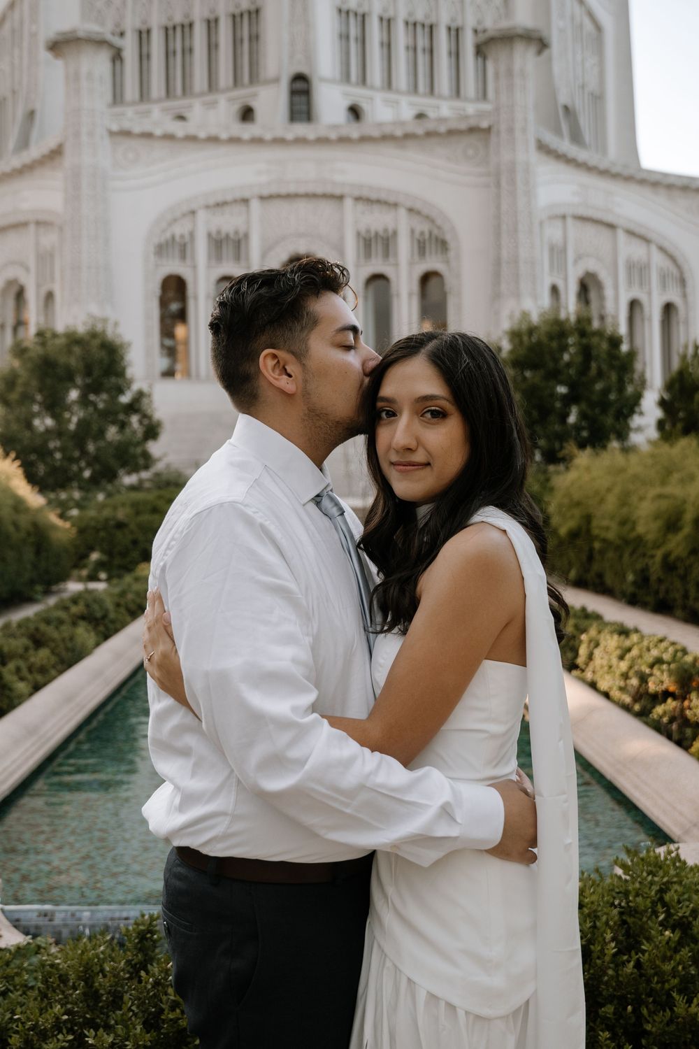 A romantic couple embraces in front of the Baha'i Temple with its ornate white architecture in the background.