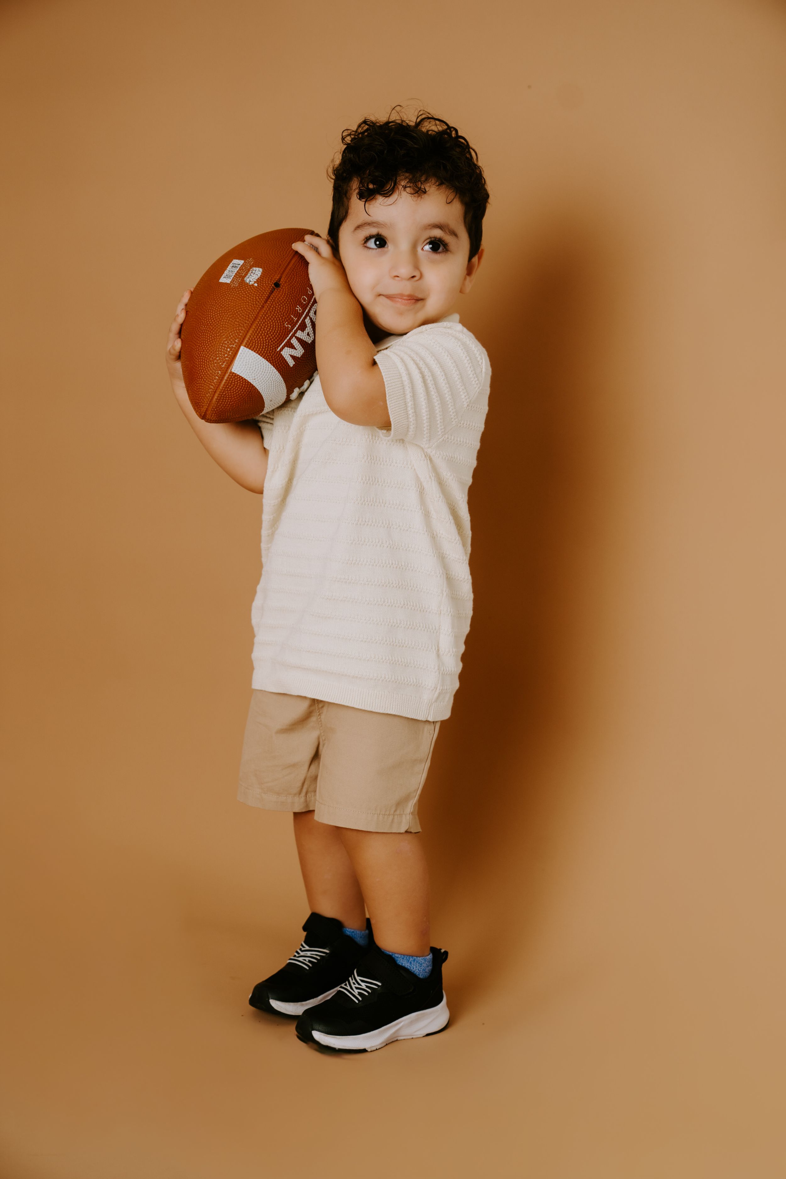 A toddler in white clothing holding a football against a brown background.