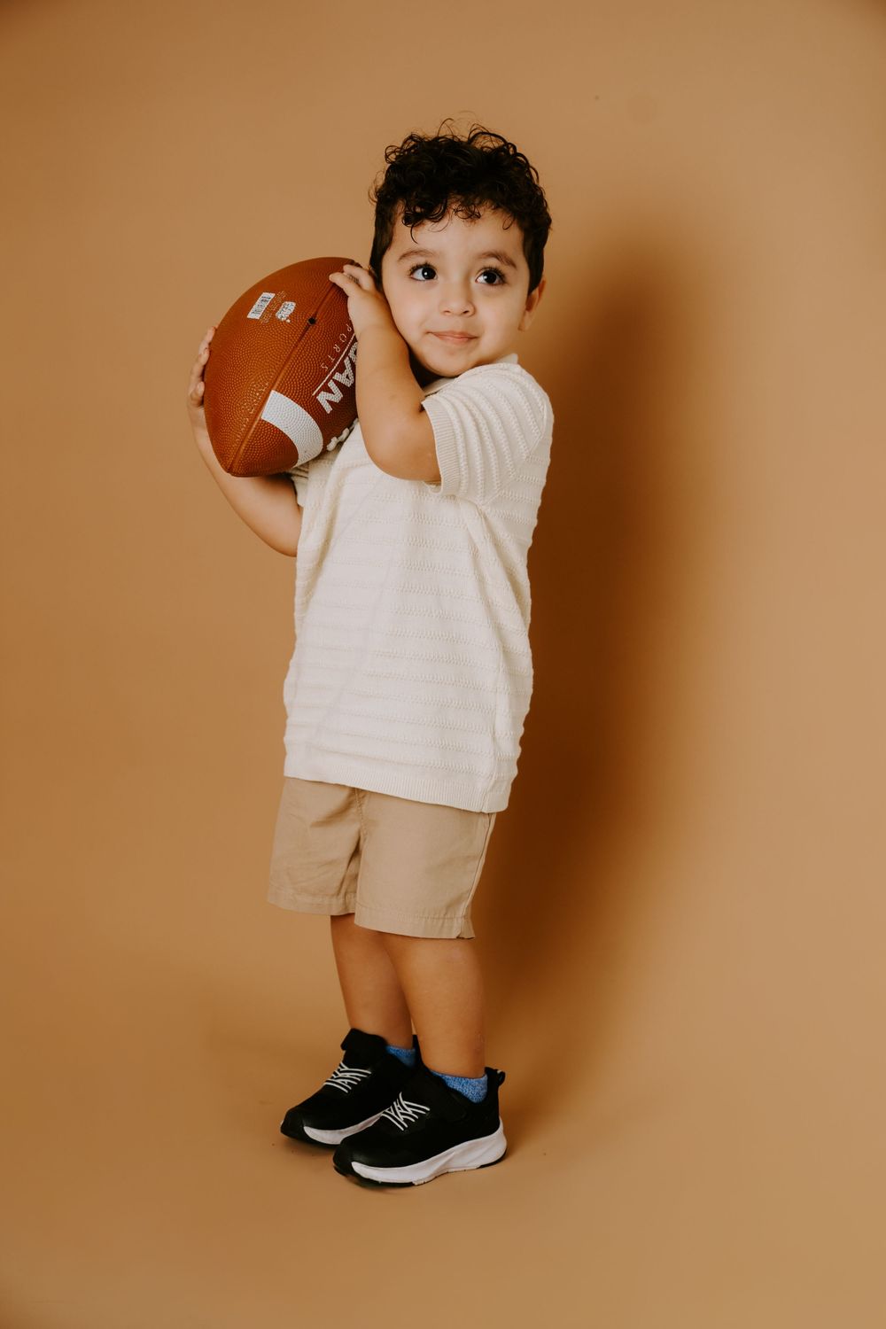 A toddler in white clothing holding a football against a brown background.