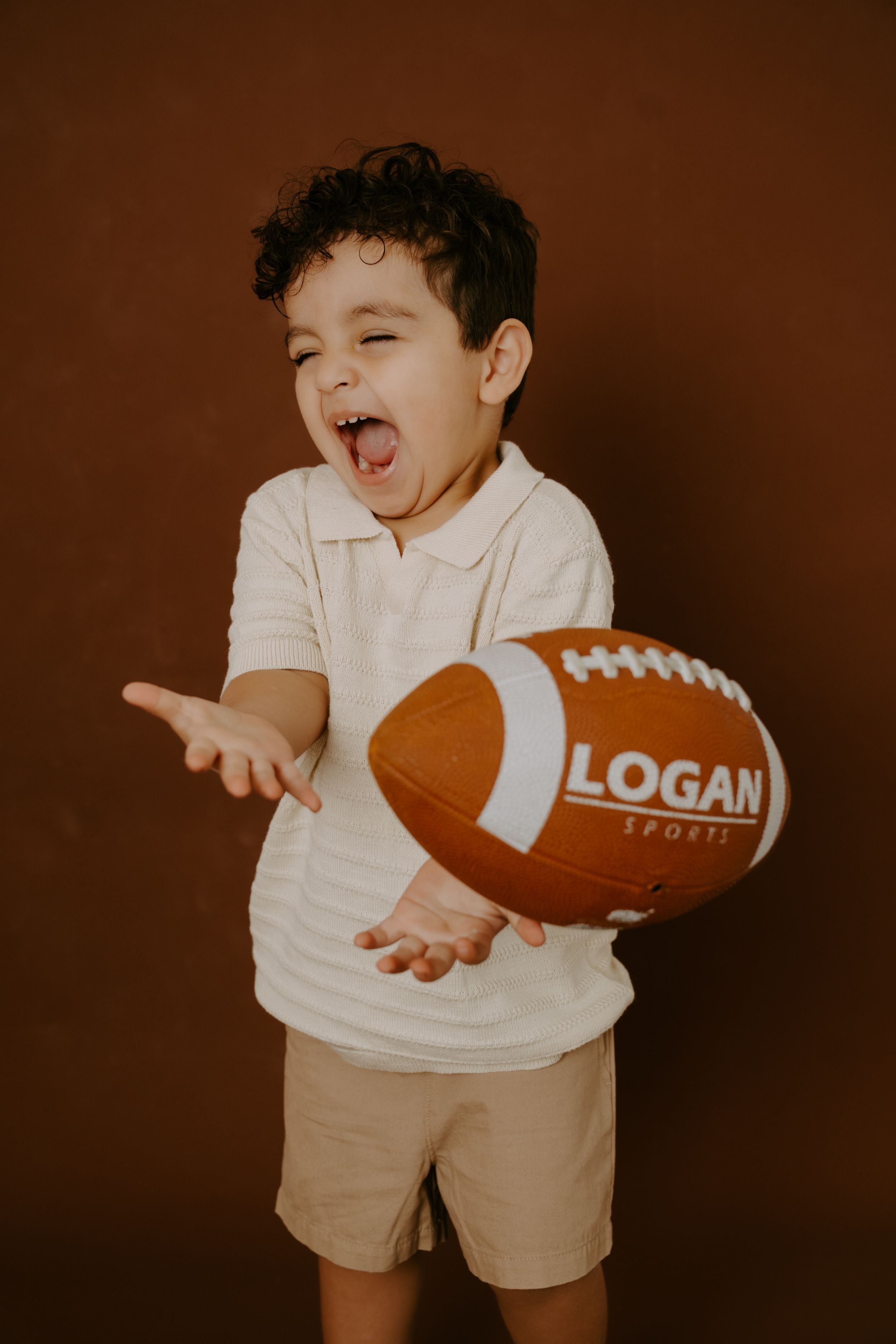 A small child in white clothing playing with a football labeled Logan against a brown backdrop.