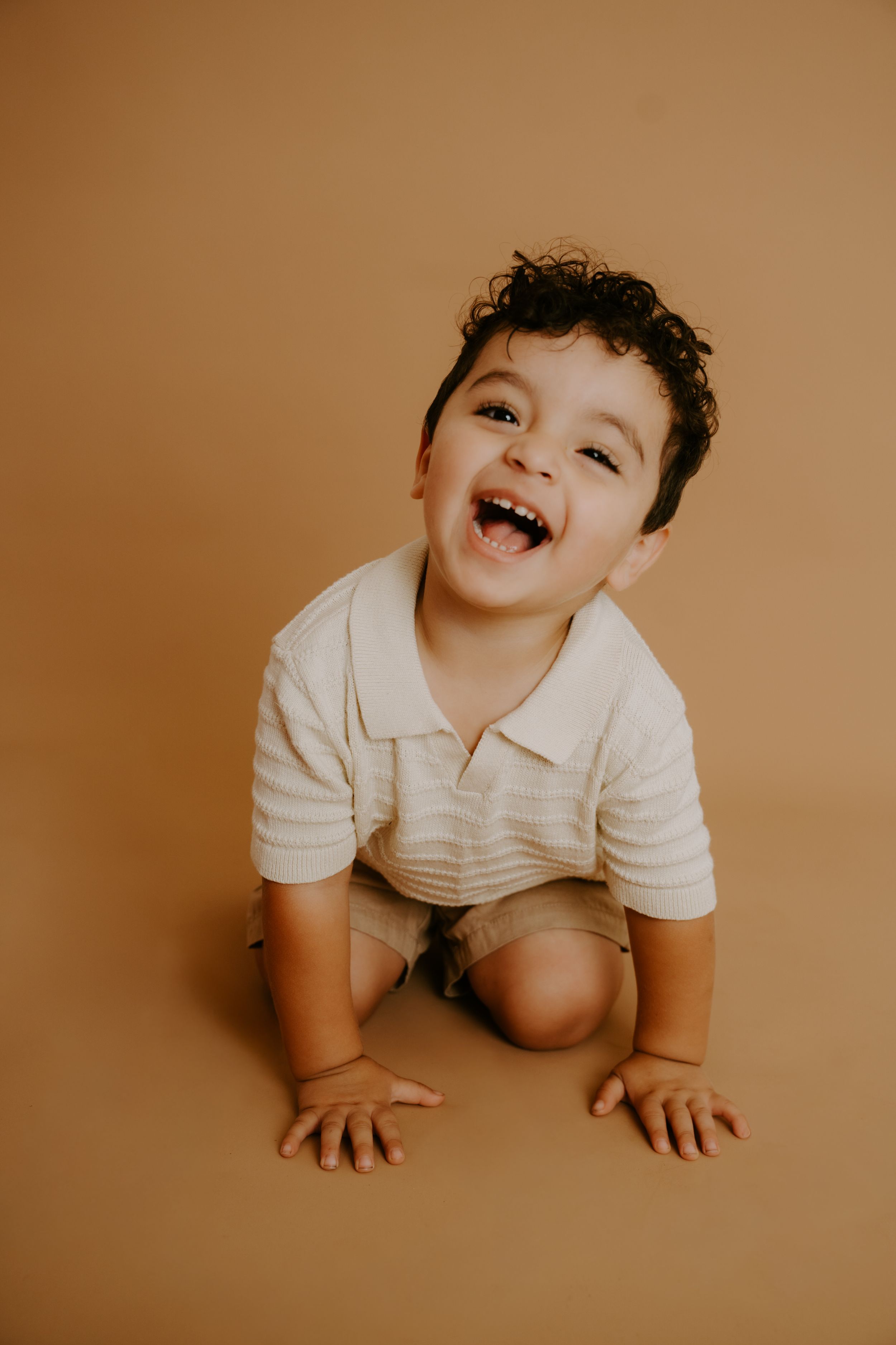 A joyful toddler in white clothing playing on a brown studio backdrop.