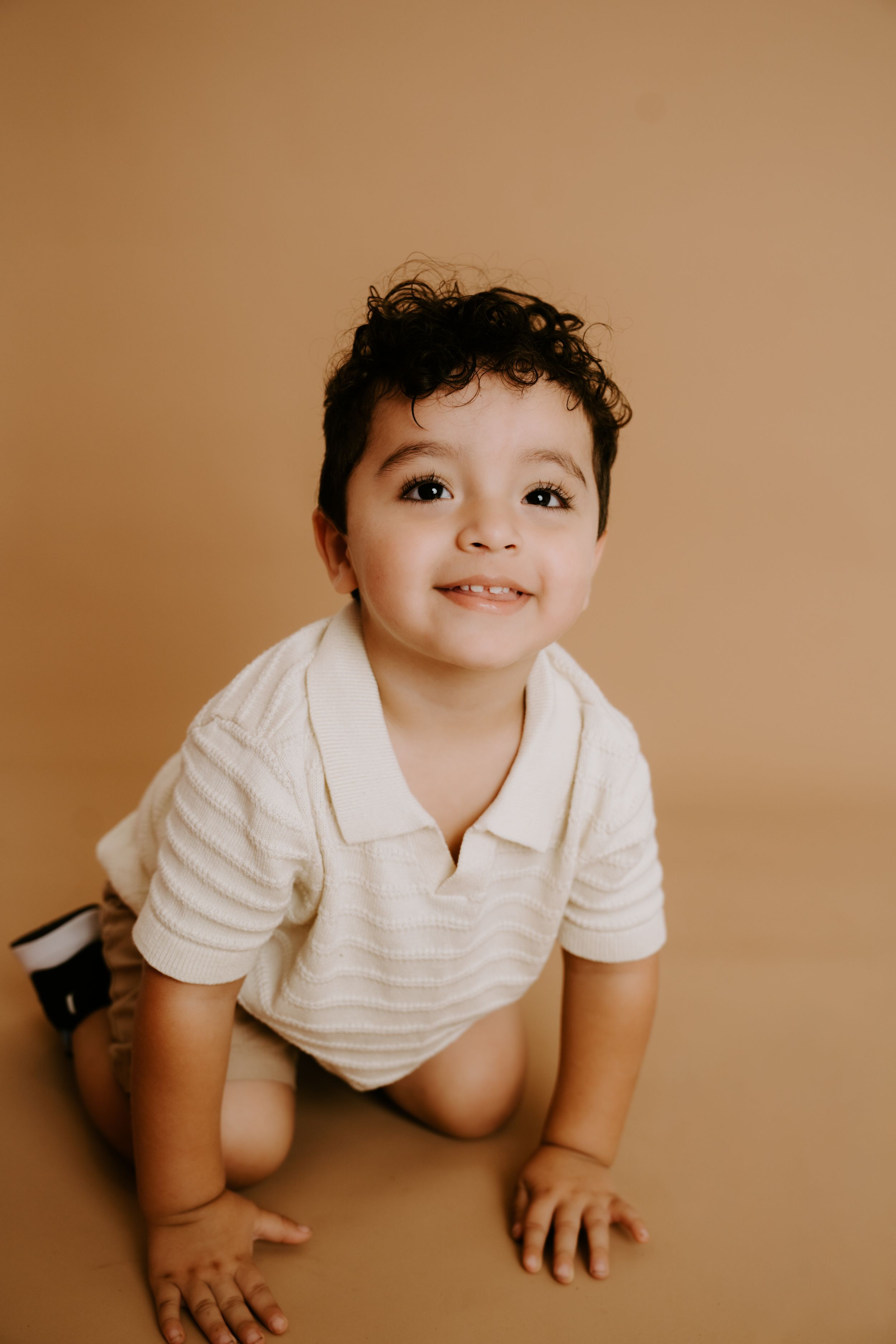 Baby in white polo shirt crawling and smiling on warm brown studio backdrop.