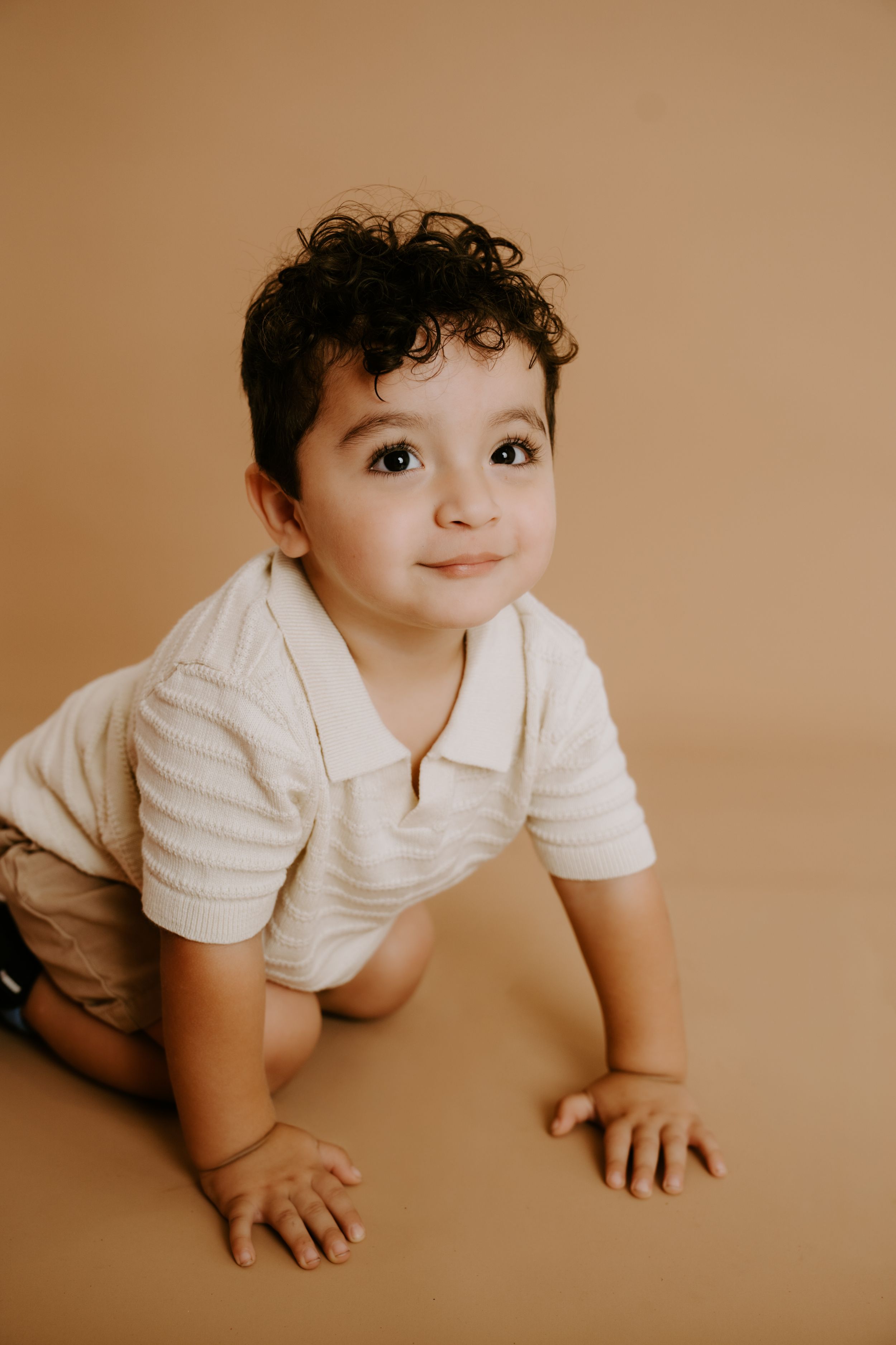 Toddler in white shirt crawling and looking up with joyful expression on brown background.