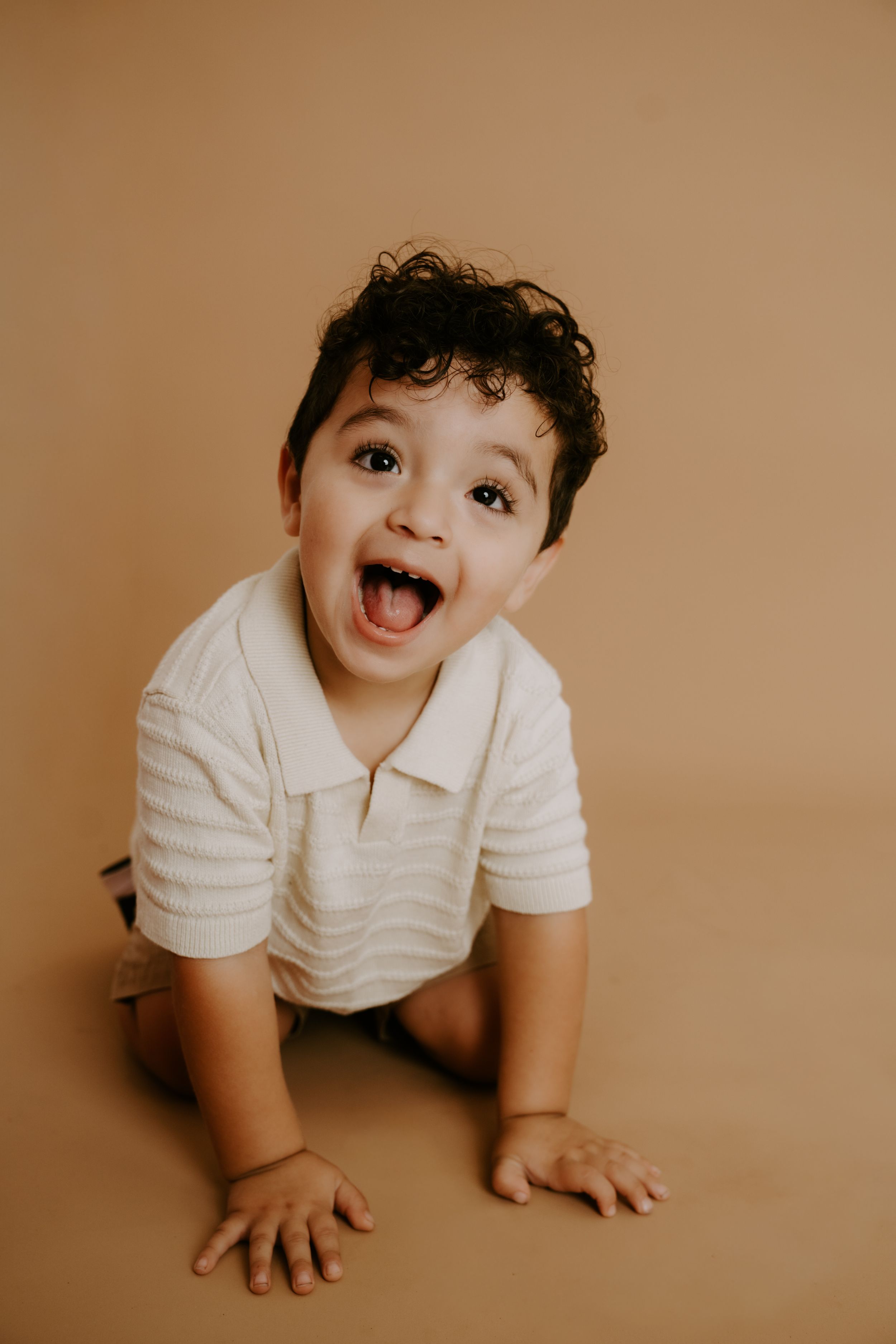 Smiling baby in white polo shirt crawling on sepia-toned studio floor.