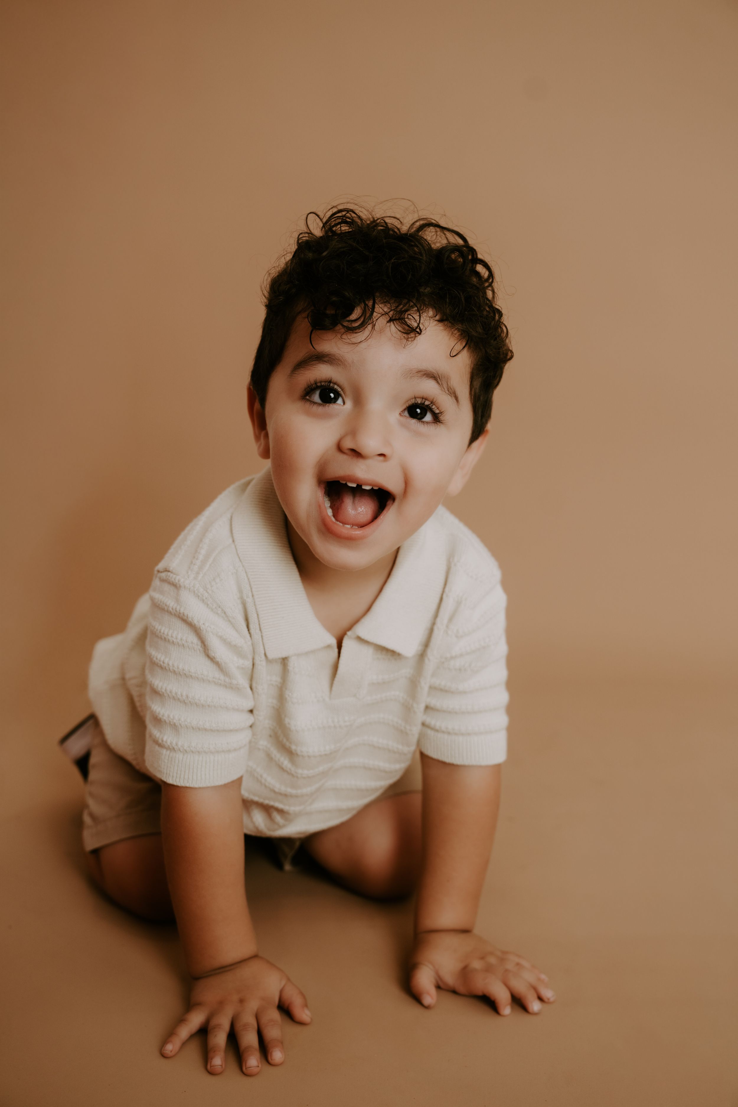 Happy baby wearing white polo shirt photographed crawling on brown studio background.
