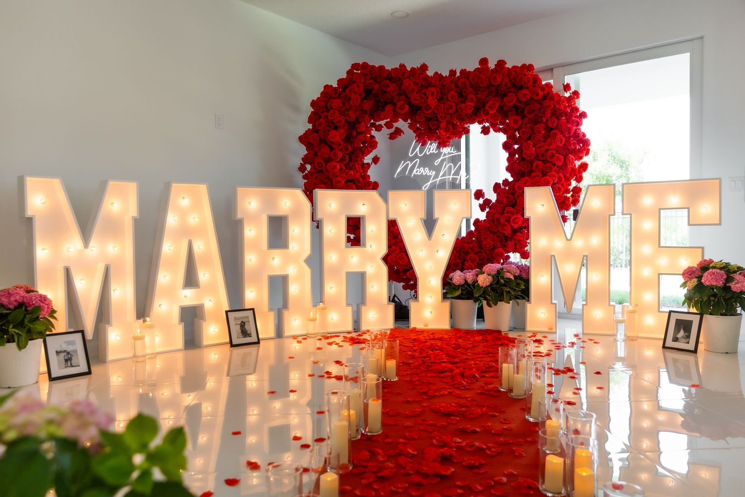 A romantic proposal setup with illuminated 'MARRY ME' letters and red rose arch decorations in a white room.