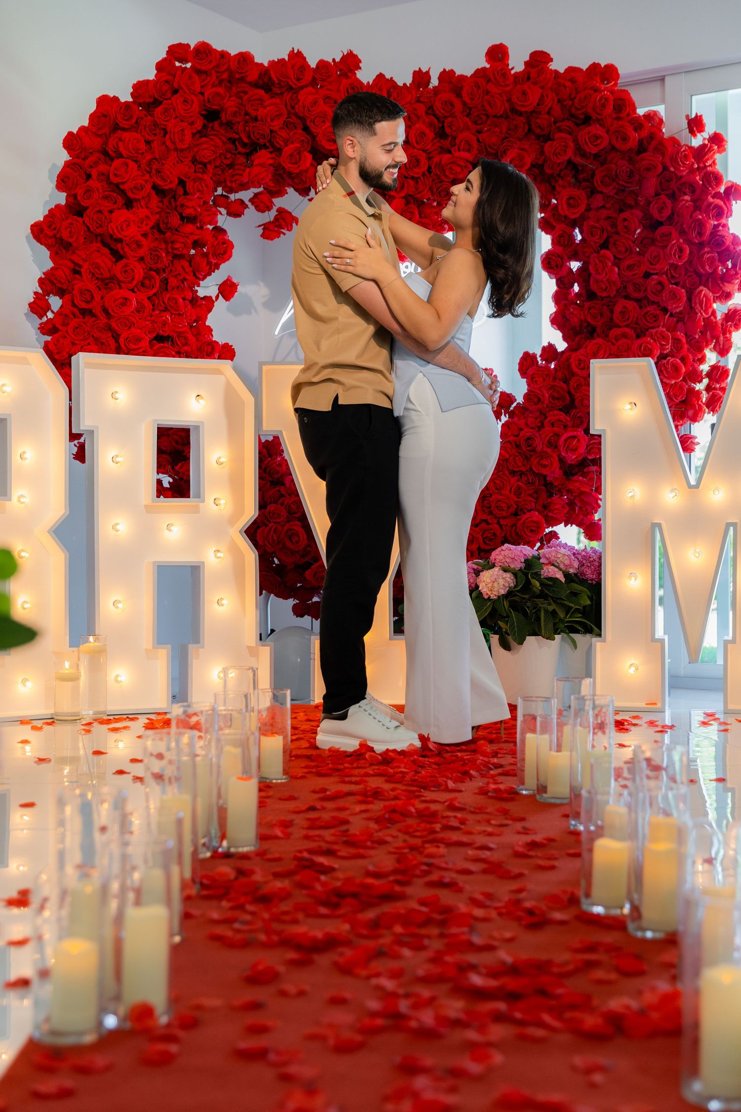 An elegant proposal scene decorated with red rose arches and illuminated white letters against a white wall.