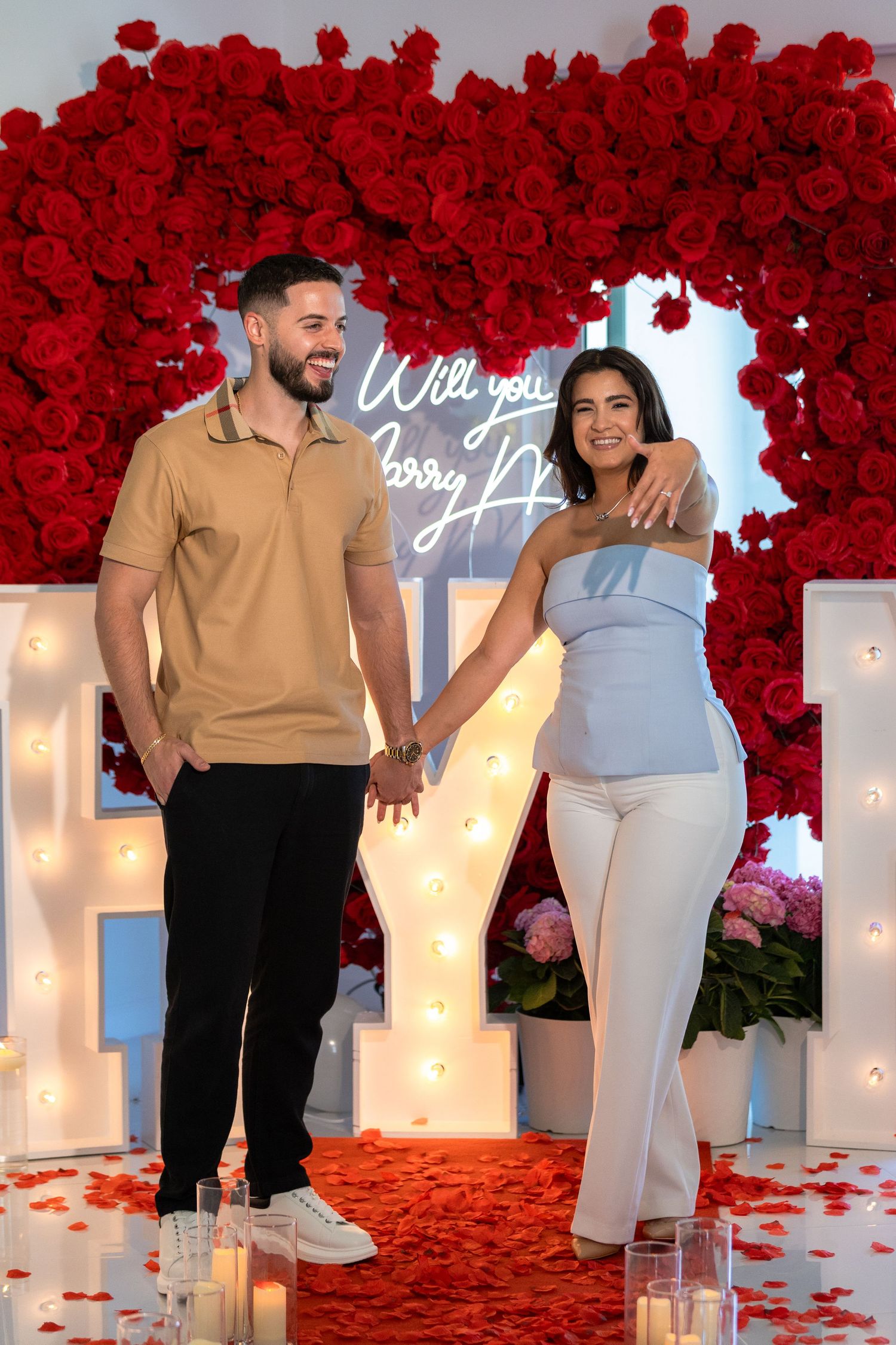 Couple poses in front of red rose wall with illuminated letters and candles for romantic engagement photos.