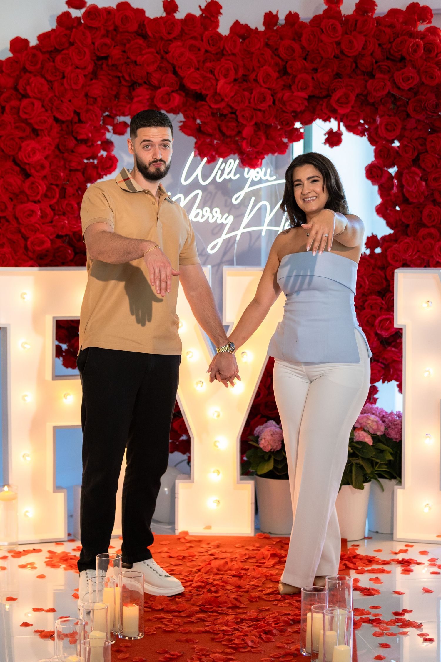 Wedding proposal scene with lit letters, red floral arch, and scattered rose petals on the floor.