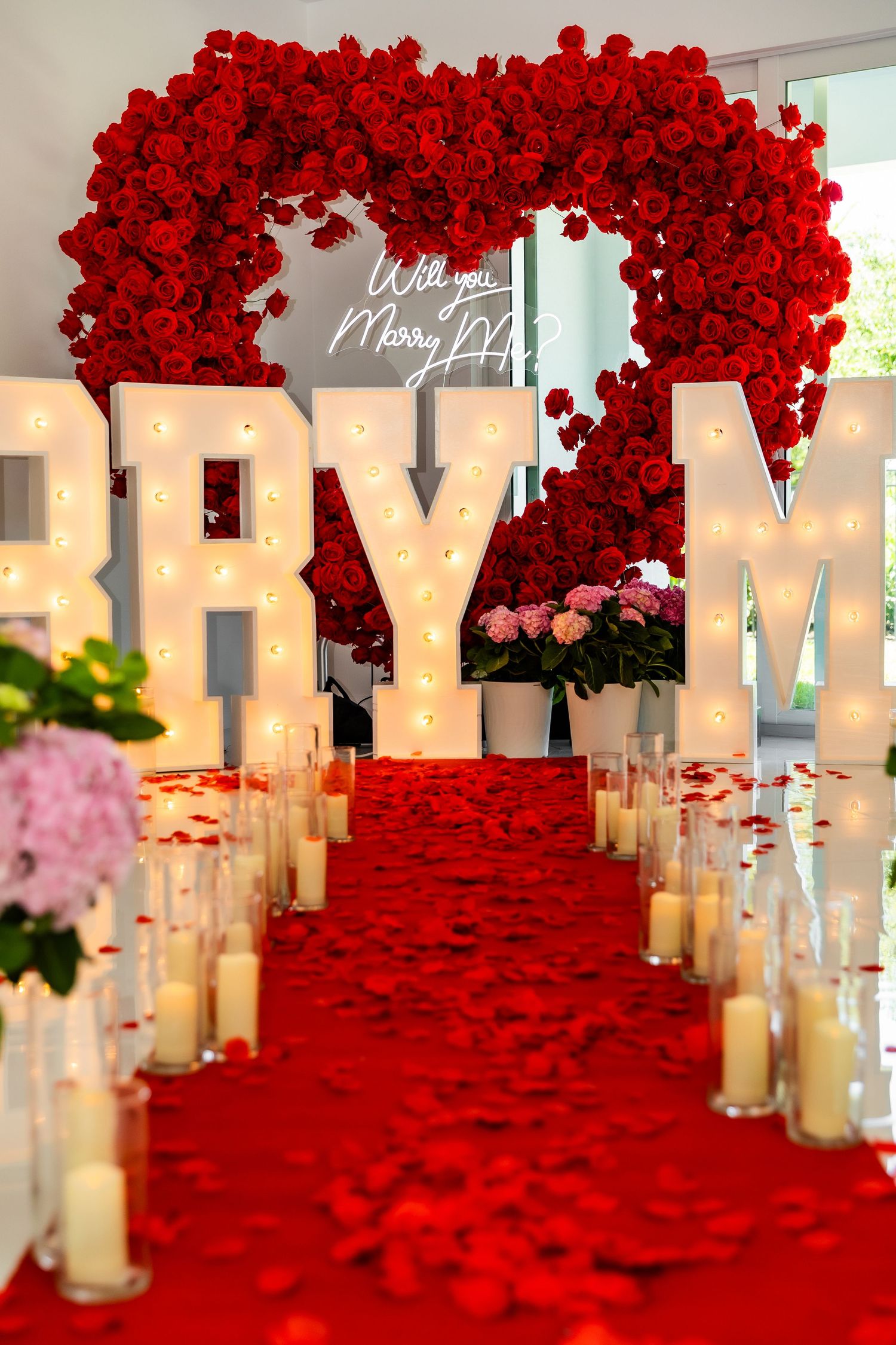 Elegant wedding proposal setup with white MARRY ME letters decorated with red roses and romantic candle-lit aisle.