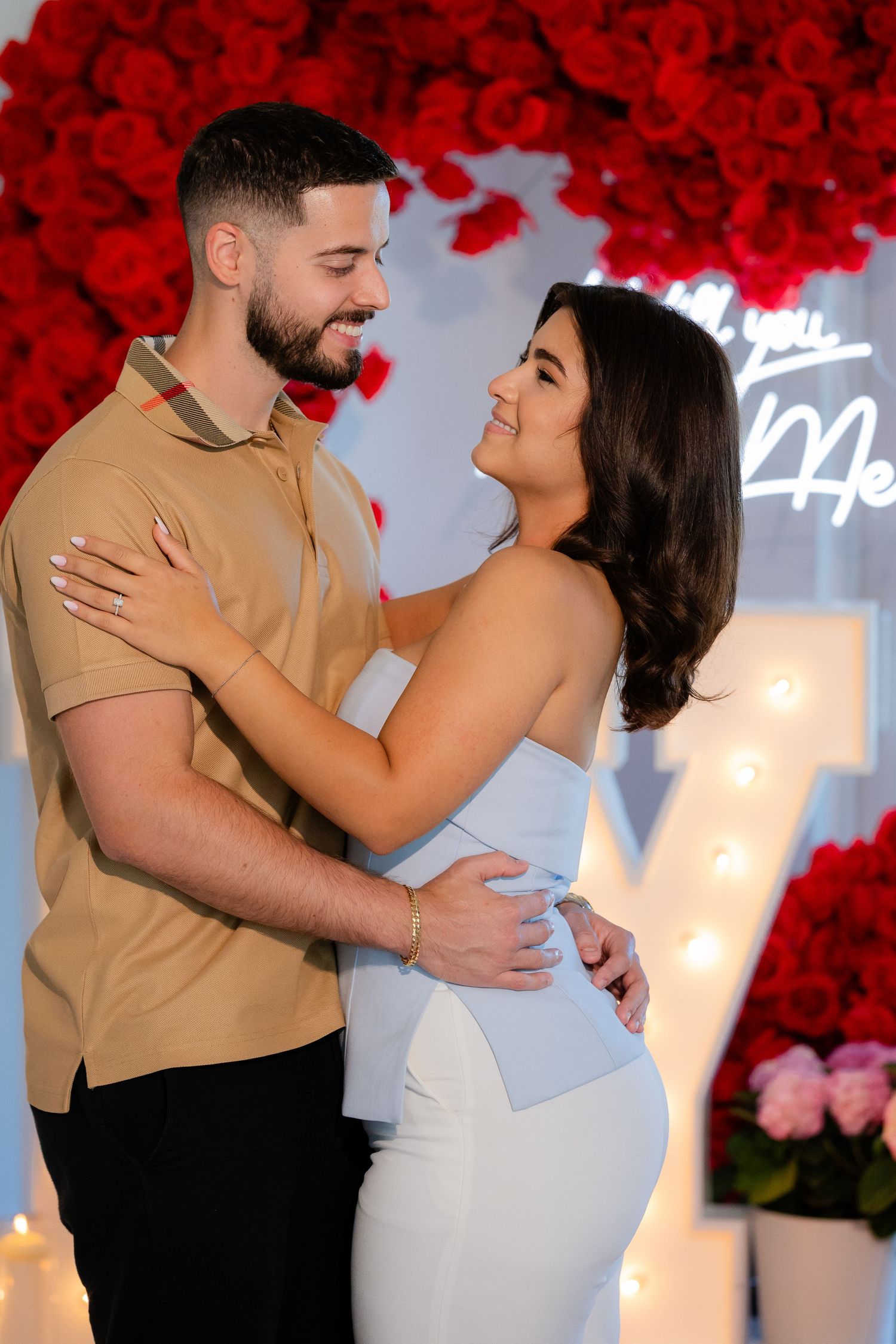 A couple poses together against a backdrop with red floral decorations during what appears to be an engagement shoot.
