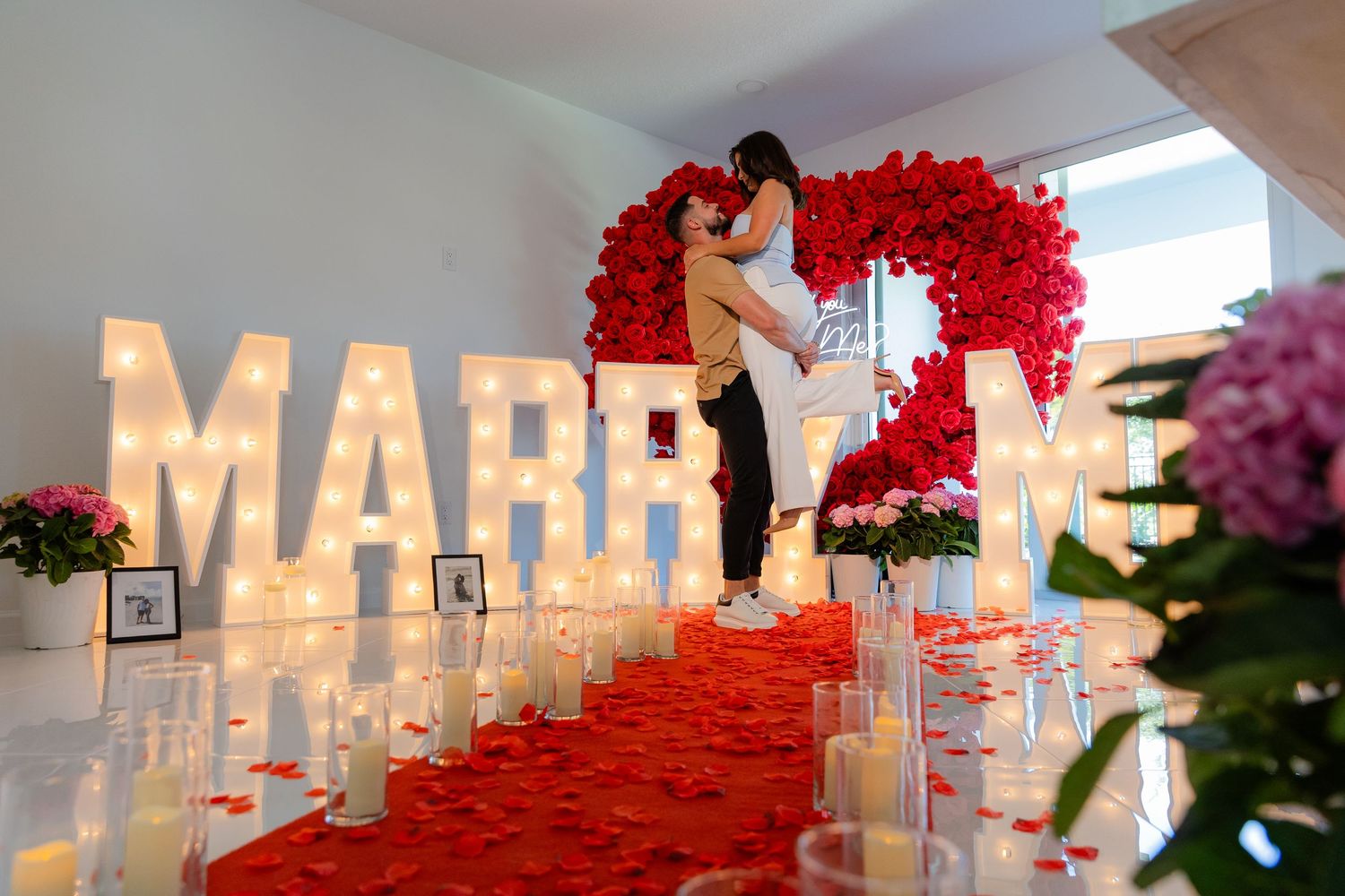 Glowing white letters spell out 'MARRY ME' surrounded by red rose petals in a romantic indoor proposal scene.