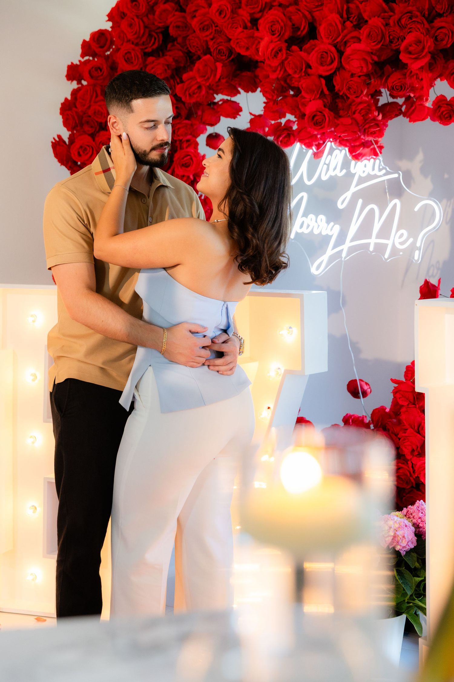 Intimate proposal moment captured under an arch of red roses with illuminated lettering in the background.