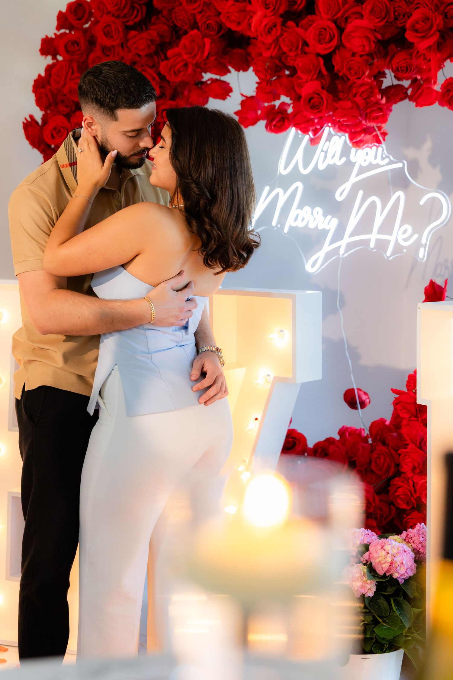 Couple sharing intimate moment under arch of red roses with lit MARRY ME sign and candles.