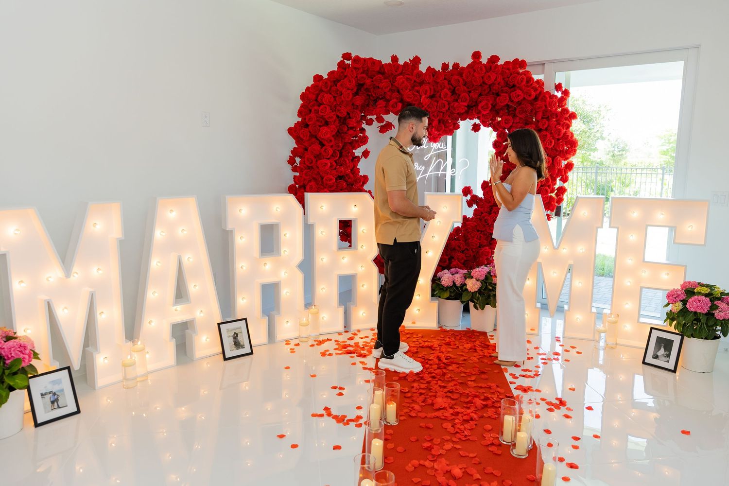 Panoramic view of a proposal setup with glowing letters, red rose petals, and candles in white interior space.