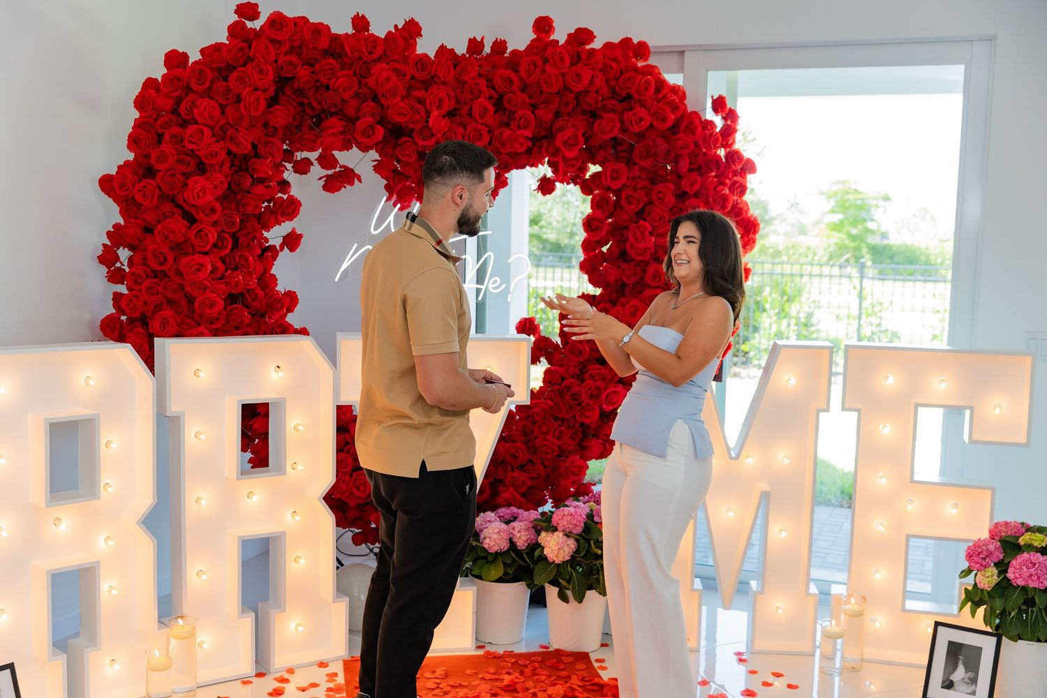 Romantic proposal setup with large illuminated letters and red floral arch during an intimate indoor celebration.