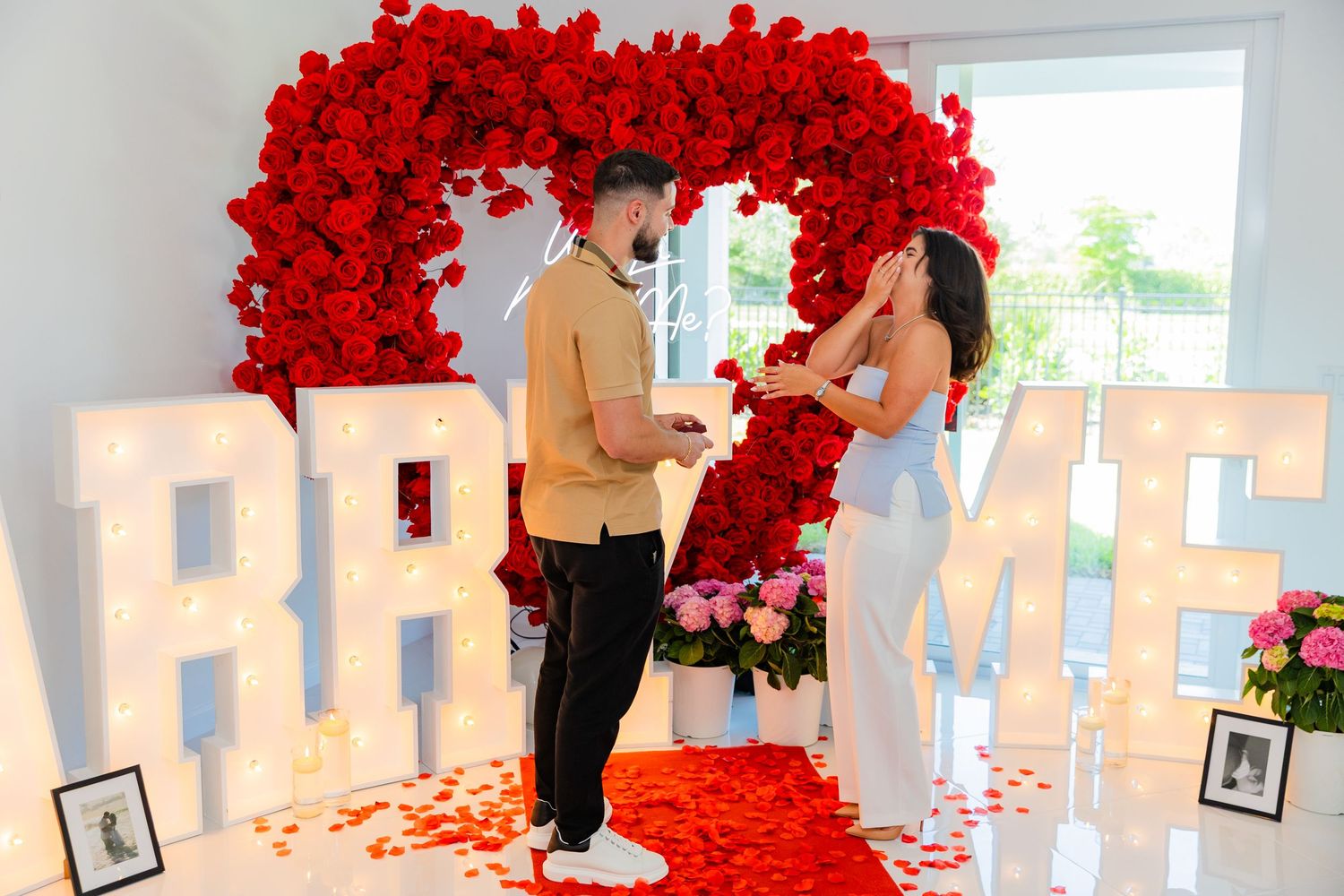 Romantic marriage proposal setup with illuminated white letters and a dramatic red rose archway overhead.