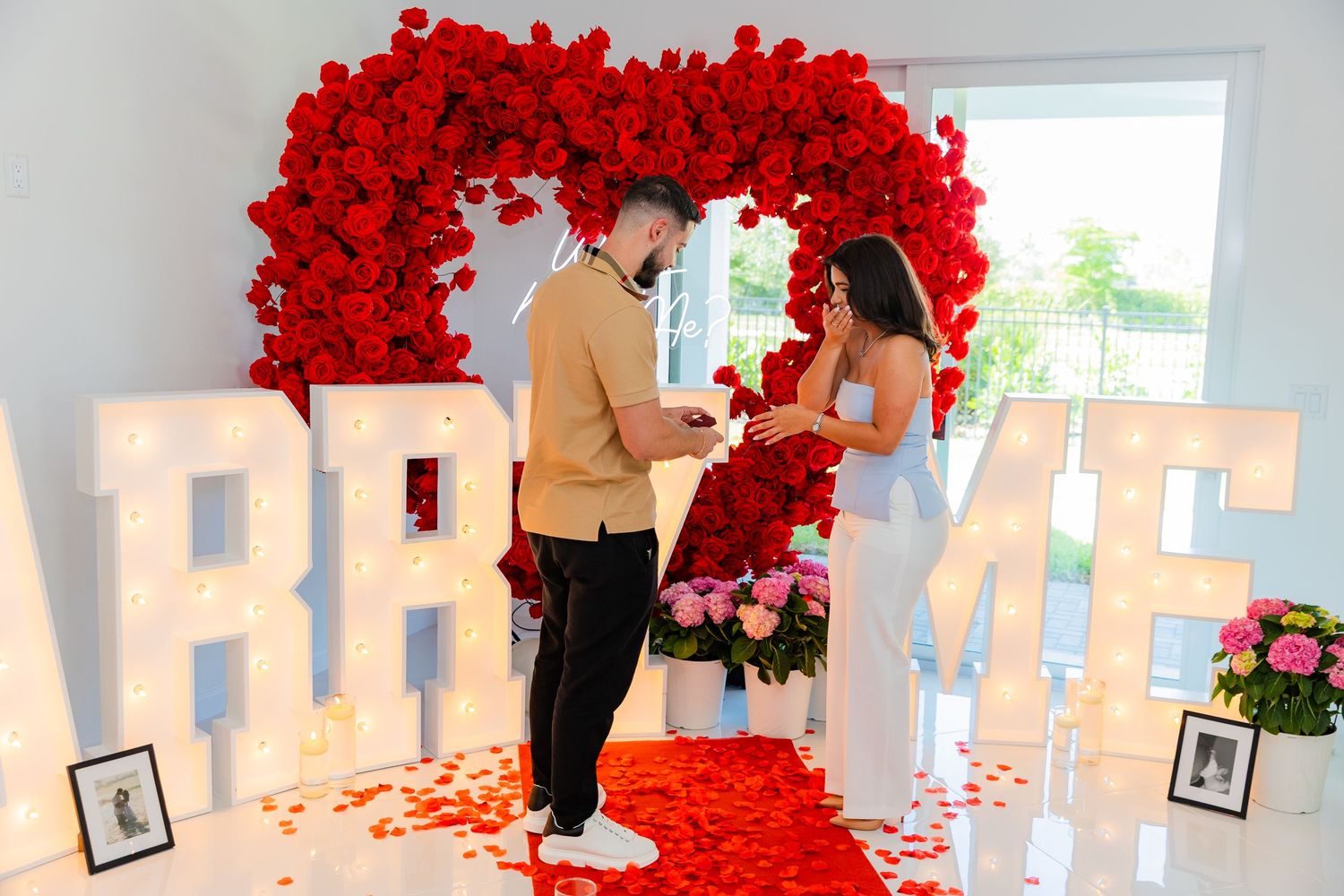 A romantic proposal setup with illuminated MARRY ME letters and a red rose arch against a white wall.