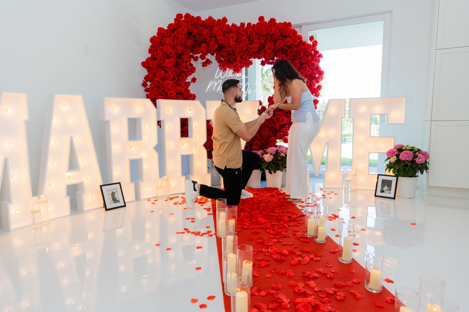 Marriage proposal scene under a bright red rose arch with glowing marquee letters.