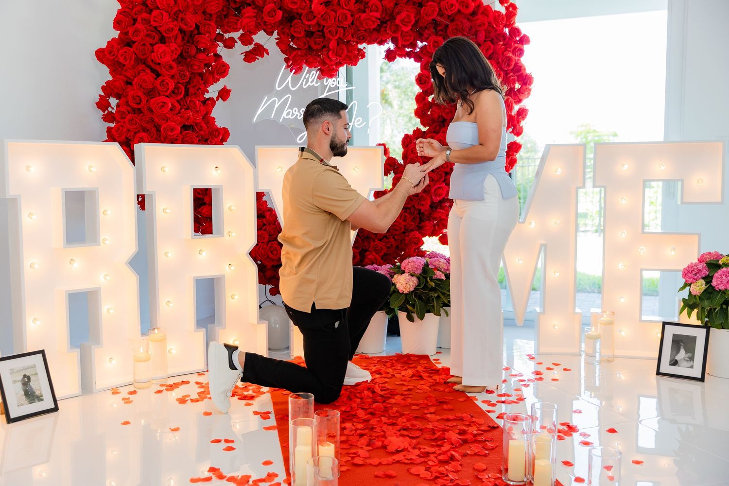 Proposal moment captured under a stunning red rose arch with illuminated MARRY ME letters in the background.
