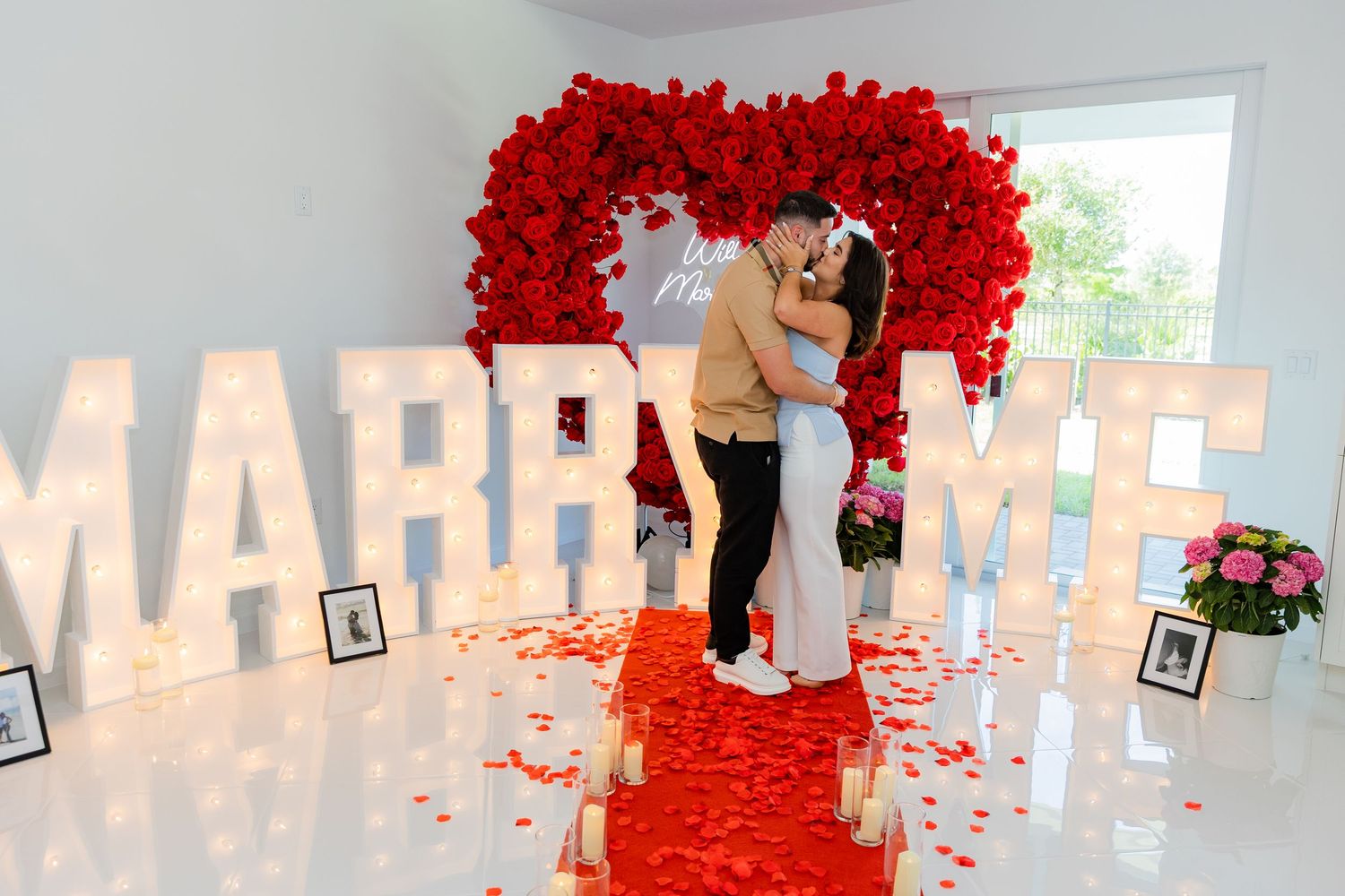 A romantic marriage proposal setup with a heart-shaped red flower arch and illuminated MARRY ME letters on a white floor.