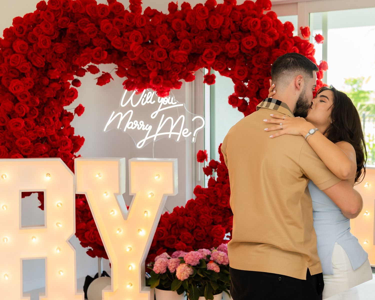 Tender moment captured during proposal with bright red rose decorations and glowing 'Marry Me' letters.