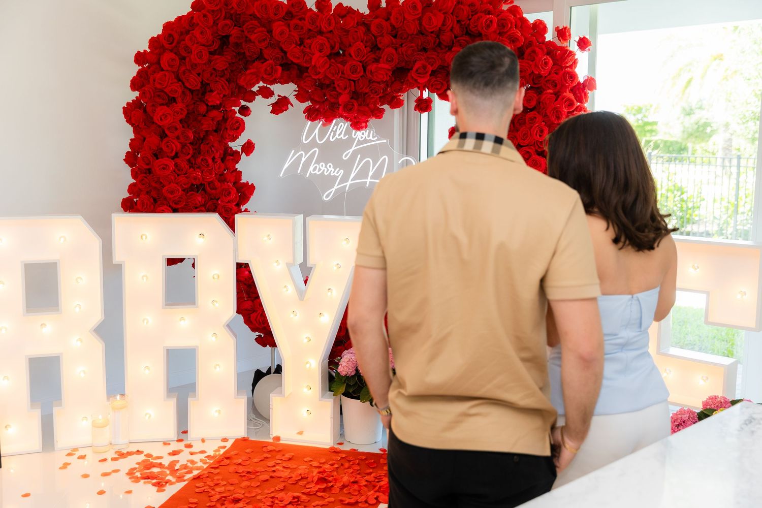 A romantic wedding proposal setup with illuminated letters and red rose petals scattered on a carpeted floor.