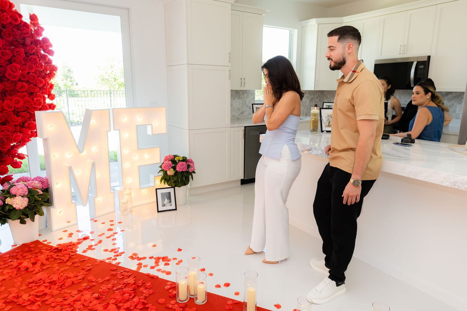 Illuminated white letters and red rose petals create a romantic marriage proposal setup in a bright indoor space.