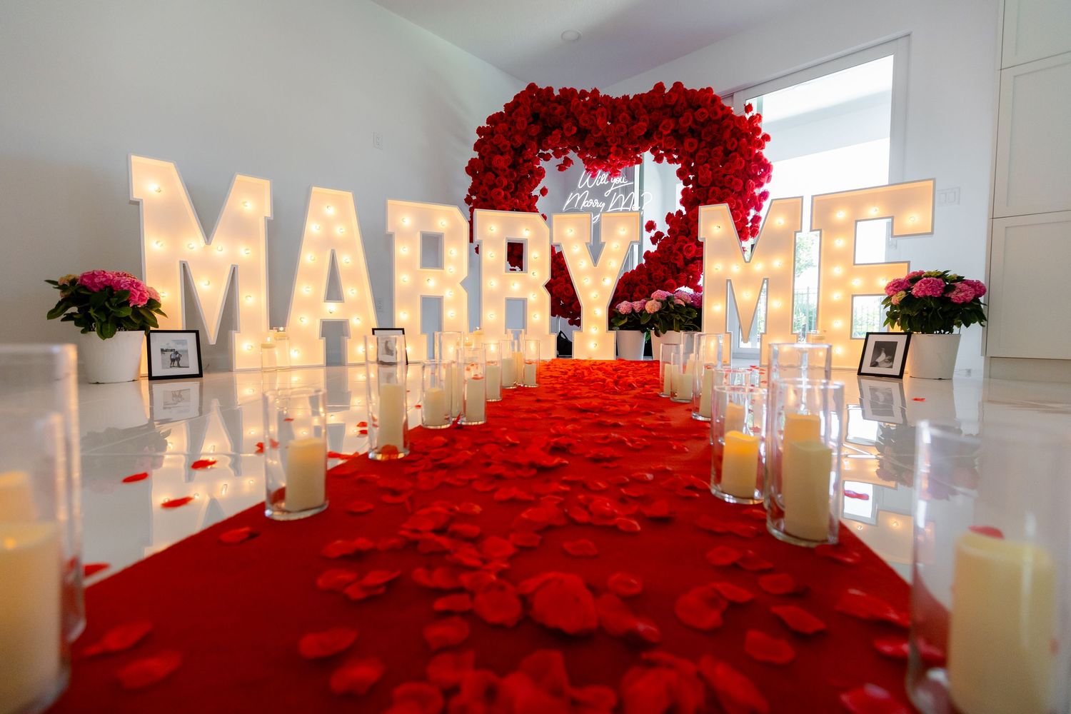 Large illuminated 'MARRY ME' sign displayed in white room with romantic red rose petals and candles on floor.