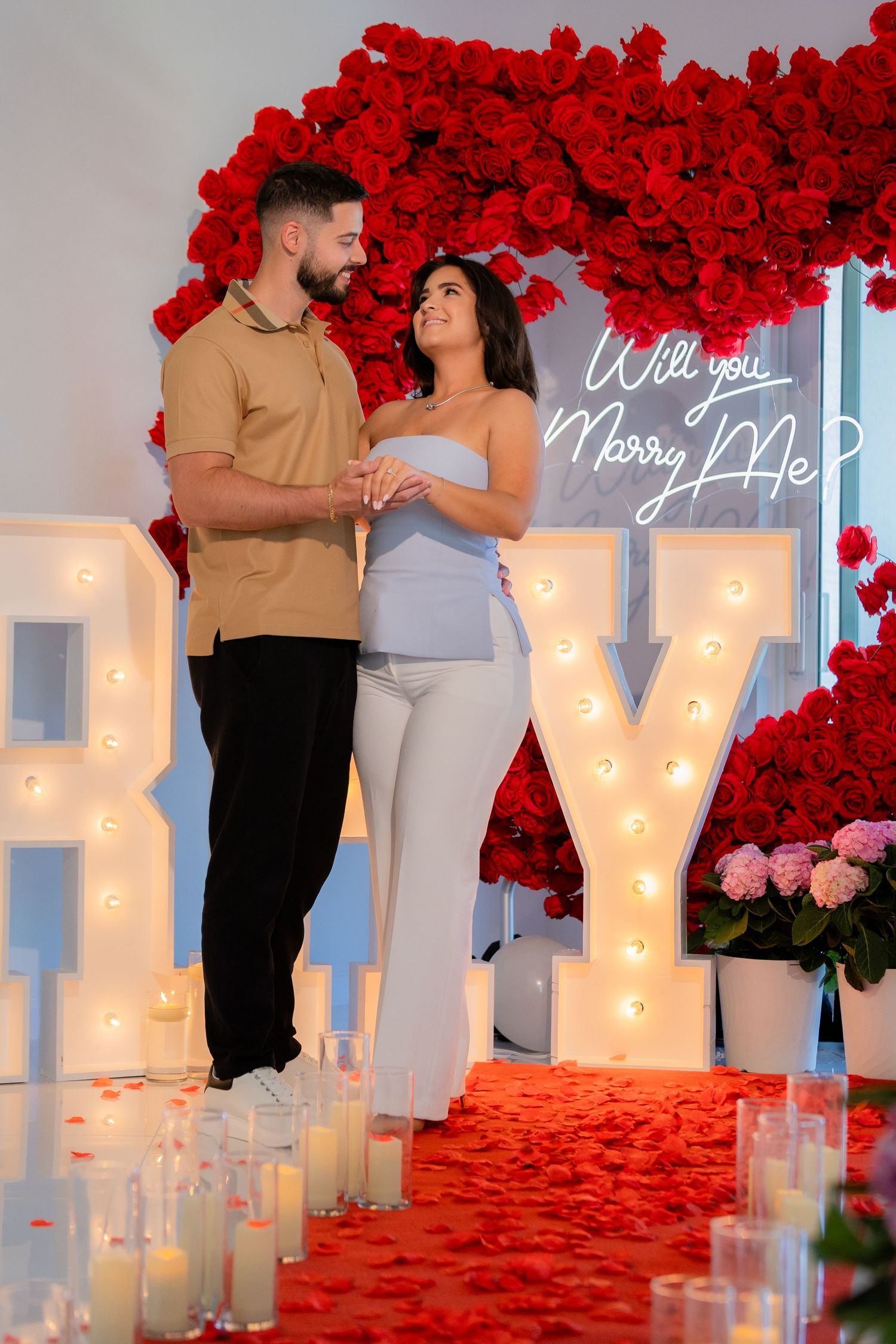 Romantic marriage proposal setup with glowing letters, red rose arch and candles lining a white aisle.