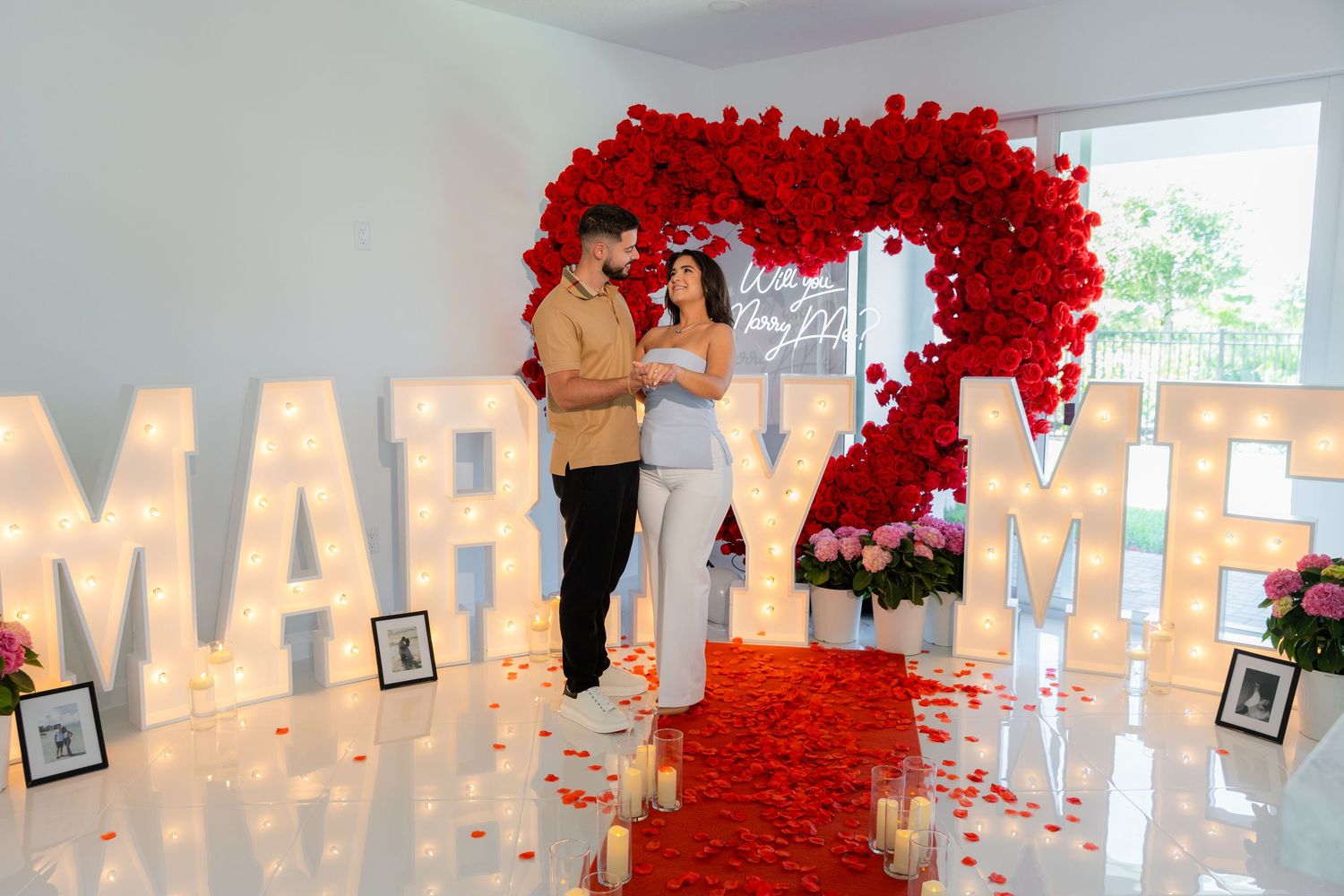 A romantic proposal setup with illuminated 'MARRY ME' letters and red rose decorations against a white wall.
